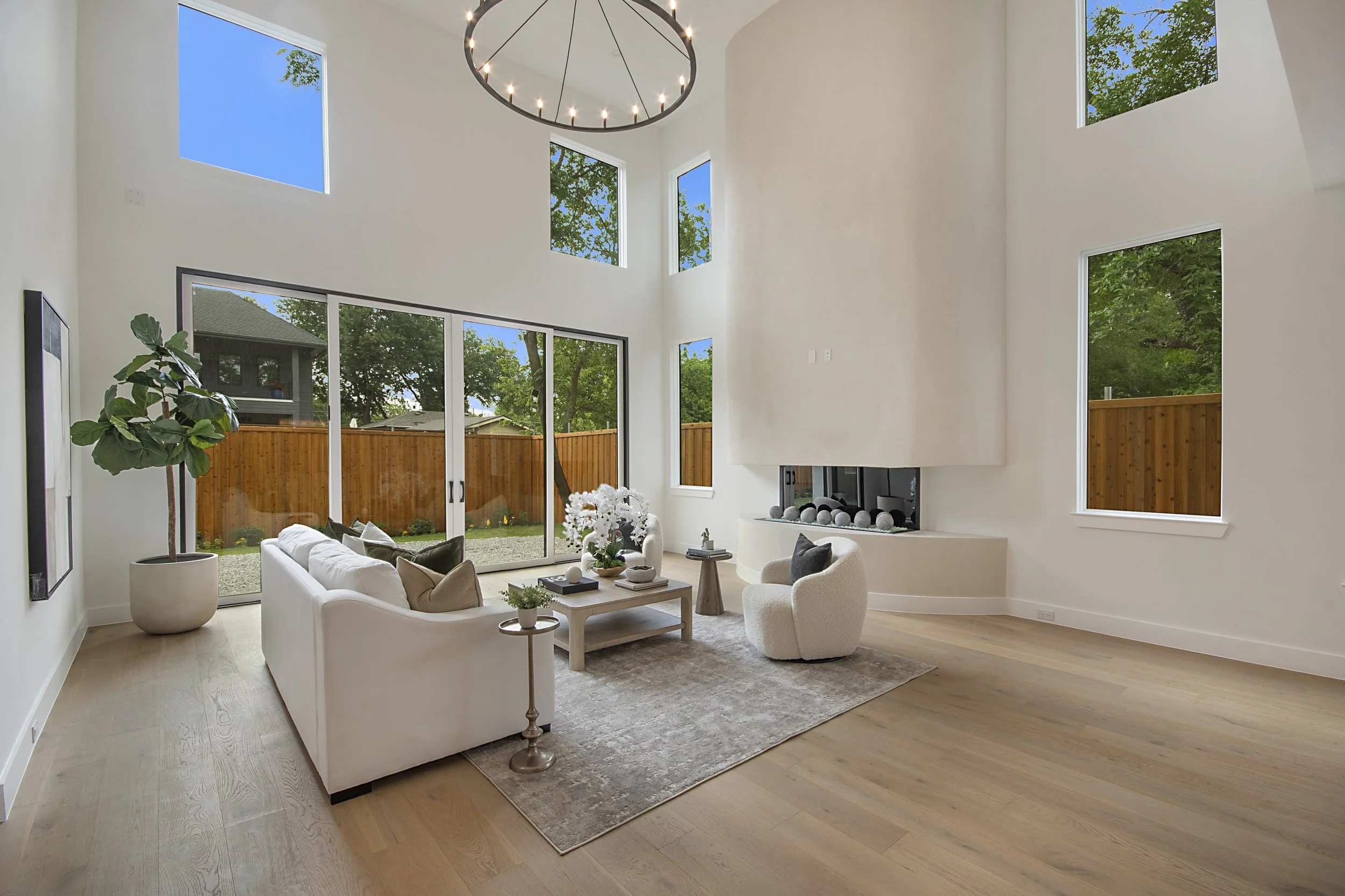 A modern living room with high ceilings and large windows showing a backyard with trees and a wooden fence. The room features white walls, a beige area rug, a white sofa with throw pillows, and a fireplace with decorative stones. There are potted pla