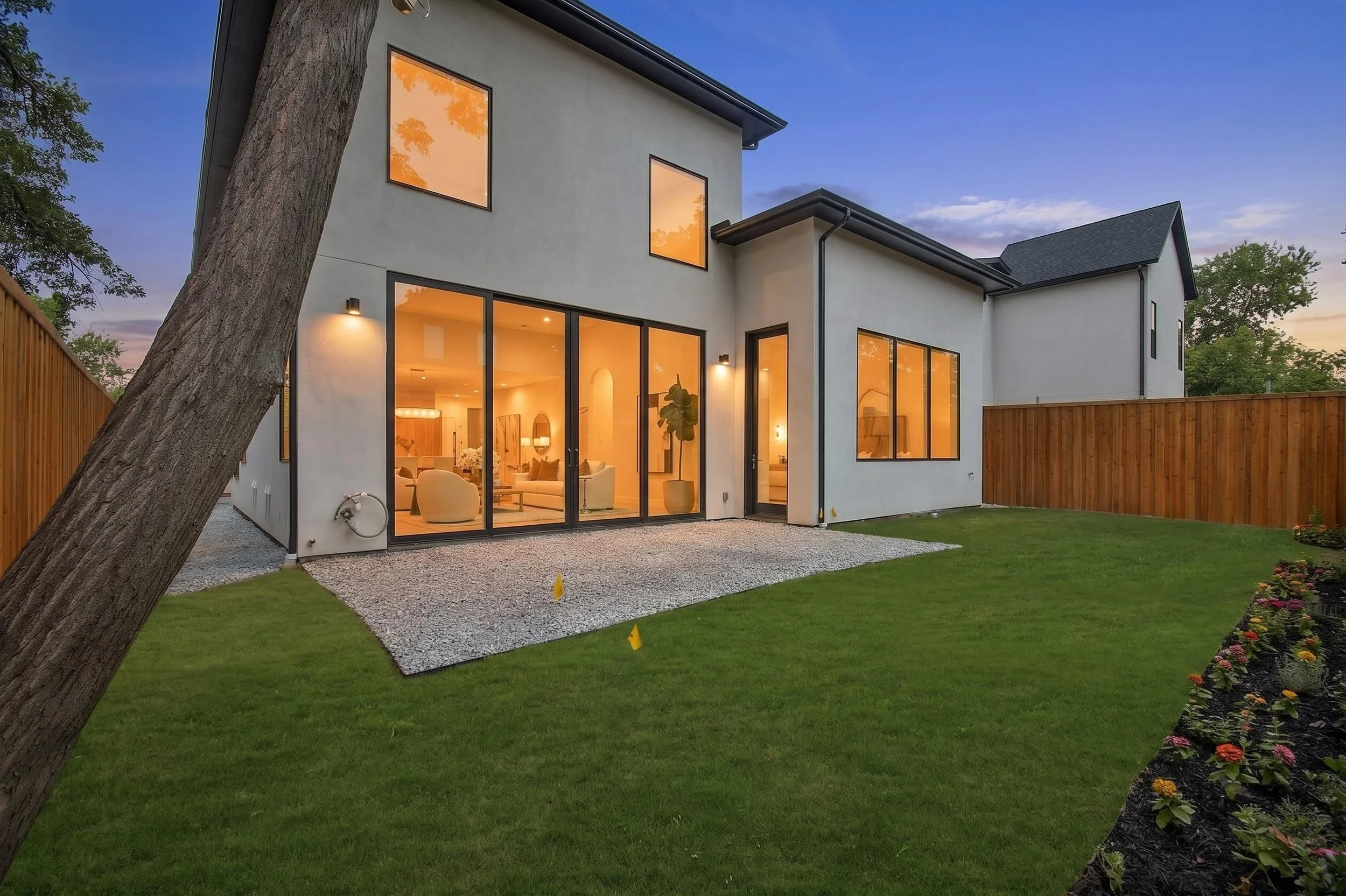 A modern two-story house with large glass windows, a wooden fence, a green lawn, and a tree in the foreground, taken at dusk with the interior lights on.