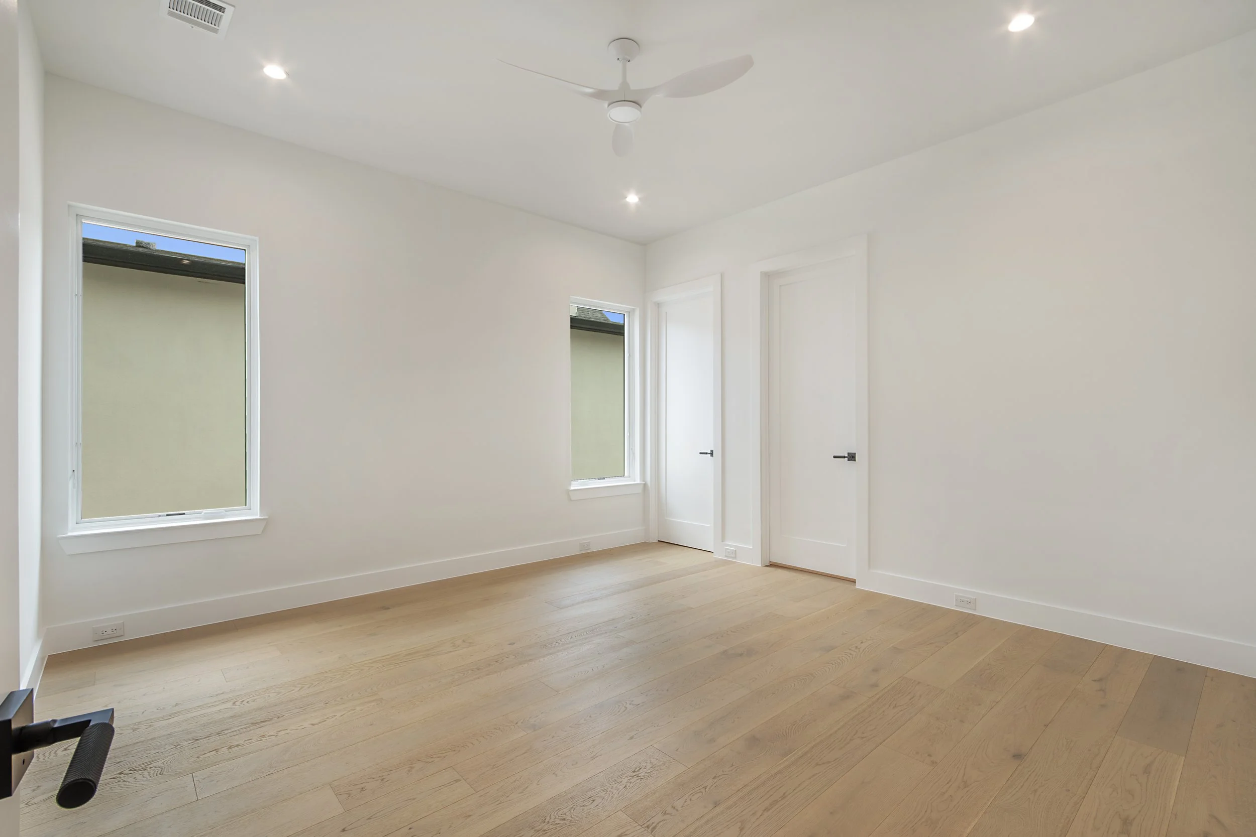 Empty room with white walls, light wood flooring, two large windows, two doors, a ceiling fan, and recessed lighting.