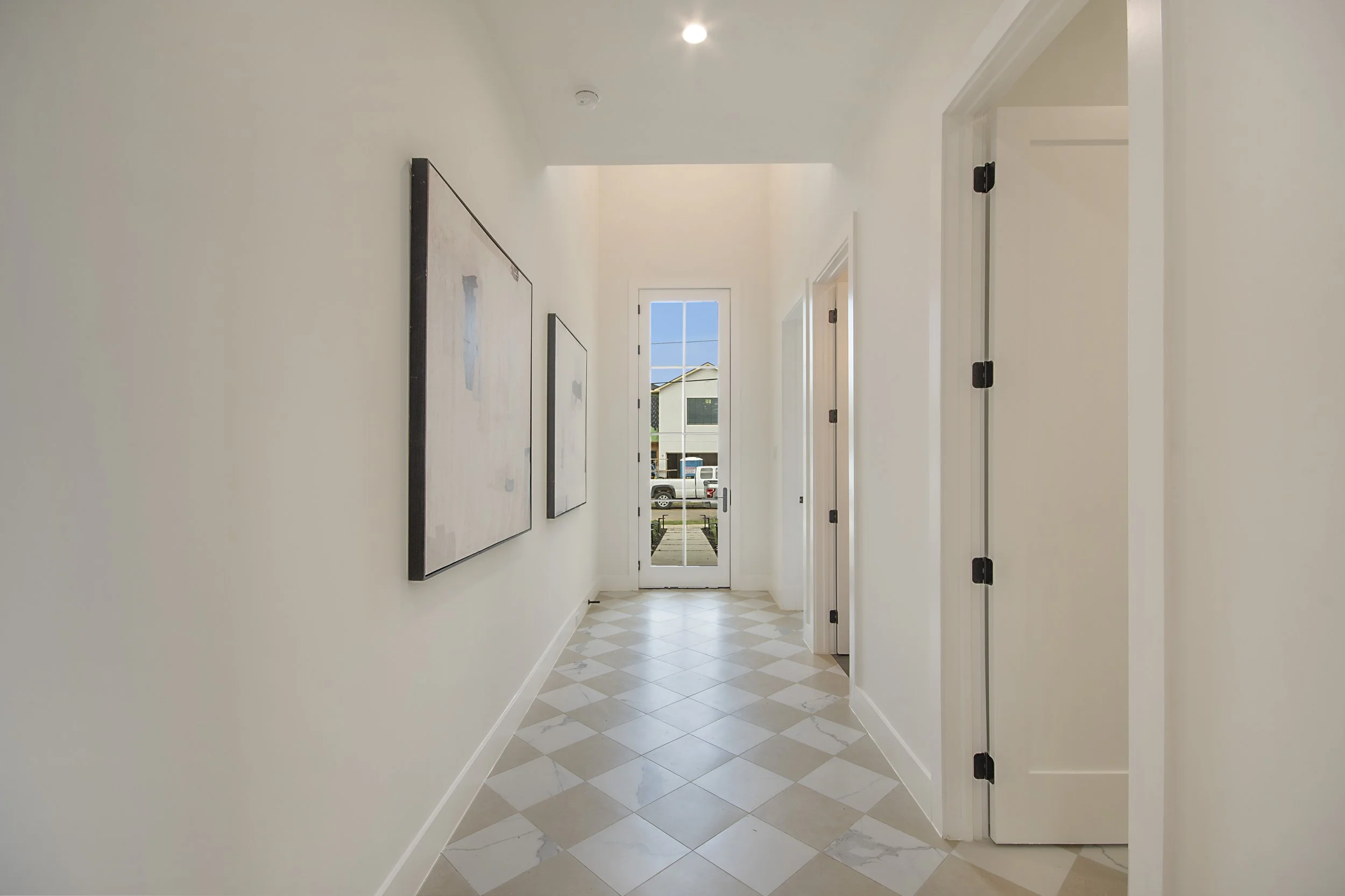 Bright hallway with white walls, light-colored tiled floor, artwork on the left wall, and a glass door at the end revealing a view of parked cars and neighboring buildings outside.