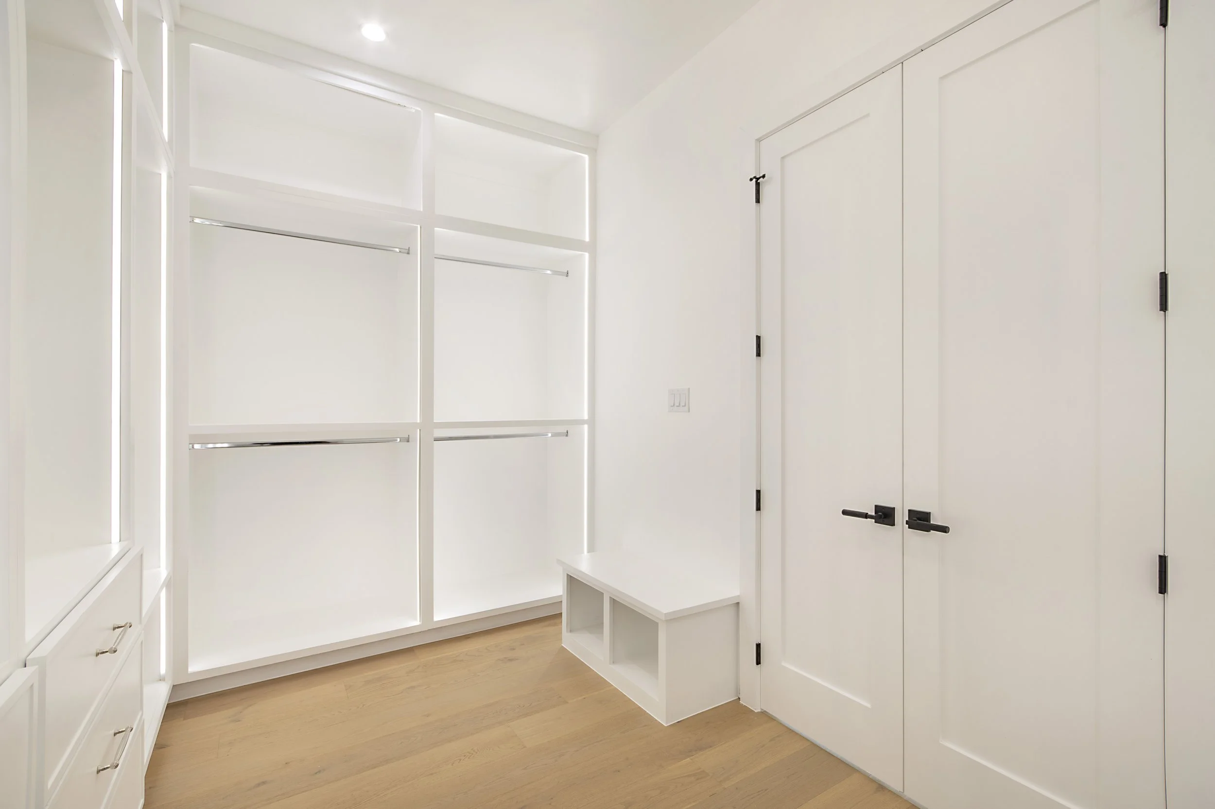 Empty white walk-in closet with built-in shelves, drawers, hanging rods, and cabinet doors, hardwood floor, ceiling light.