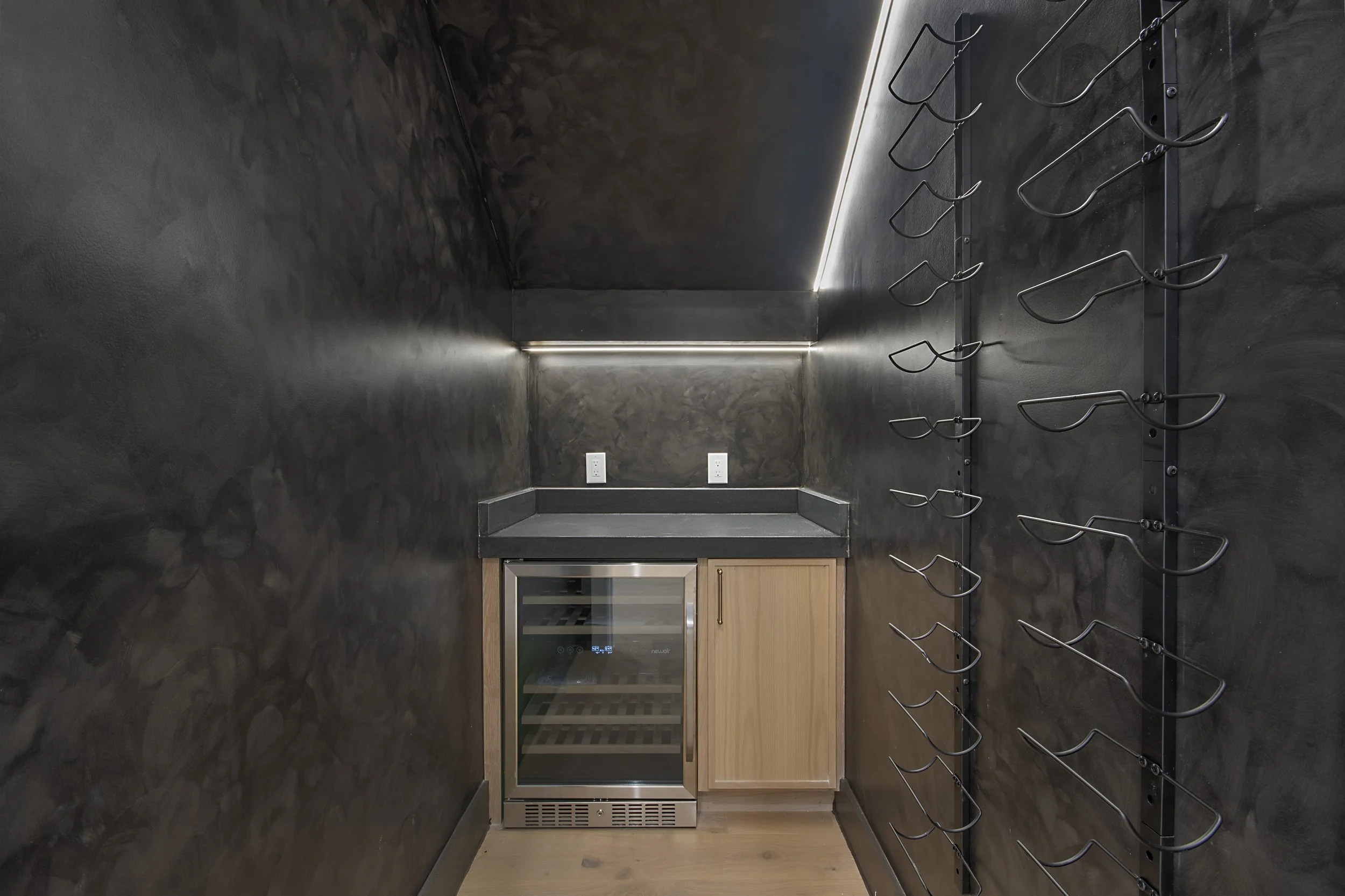 Small kitchen with dark walls, a wine cooler, light wood cabinets, and a wine rack on the right wall with empty metal slots for bottles.