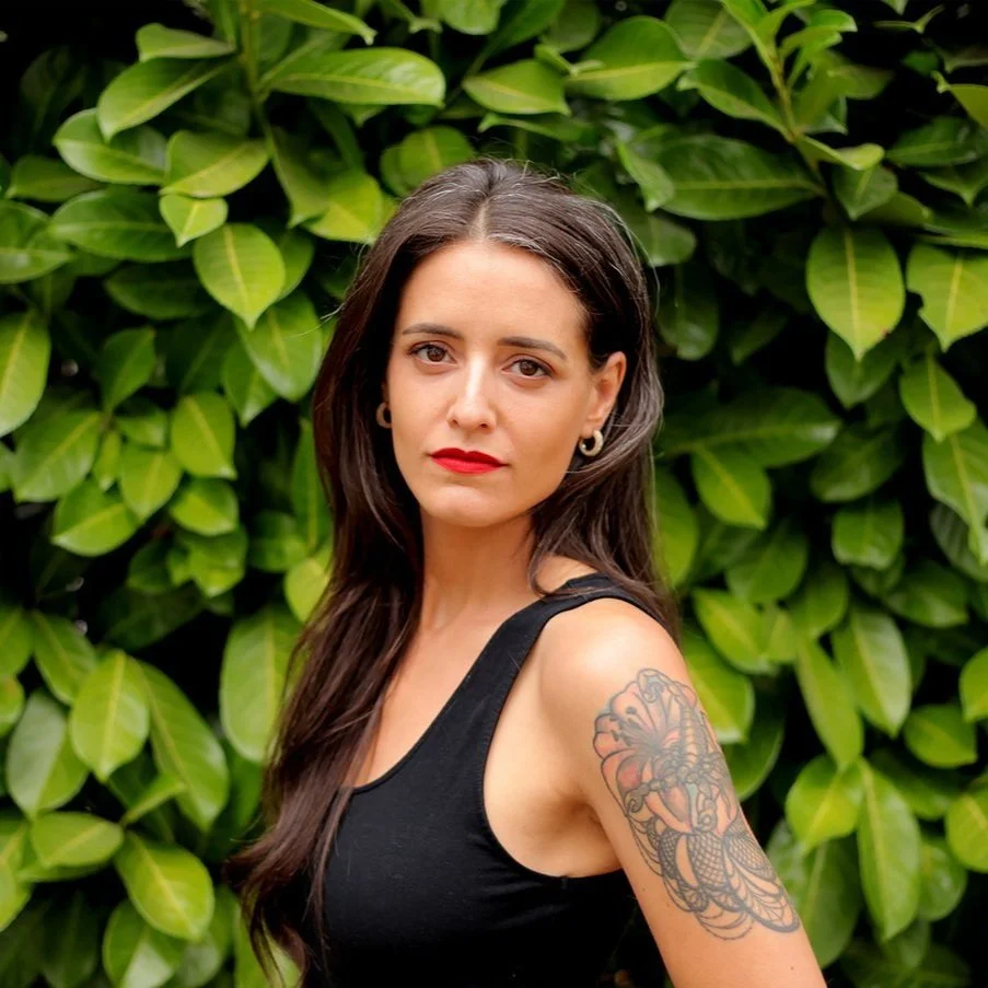 Woman with a tattoo on her upper arm standing in front of green leaves.