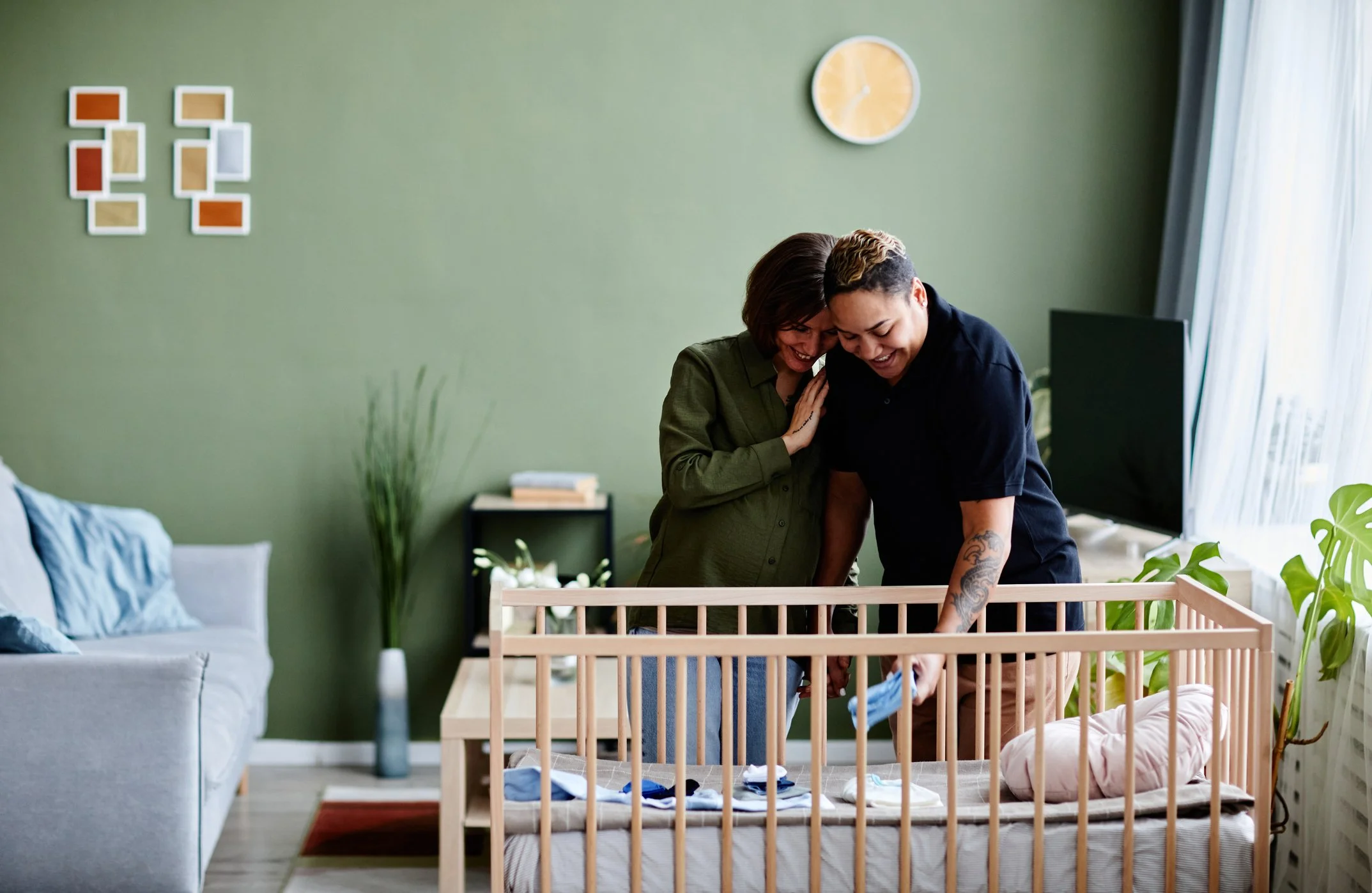Two women, one with short hair and one with longer hair, are smiling and leaning close to each other over a crib in a living room, engaging in a joyful moment.