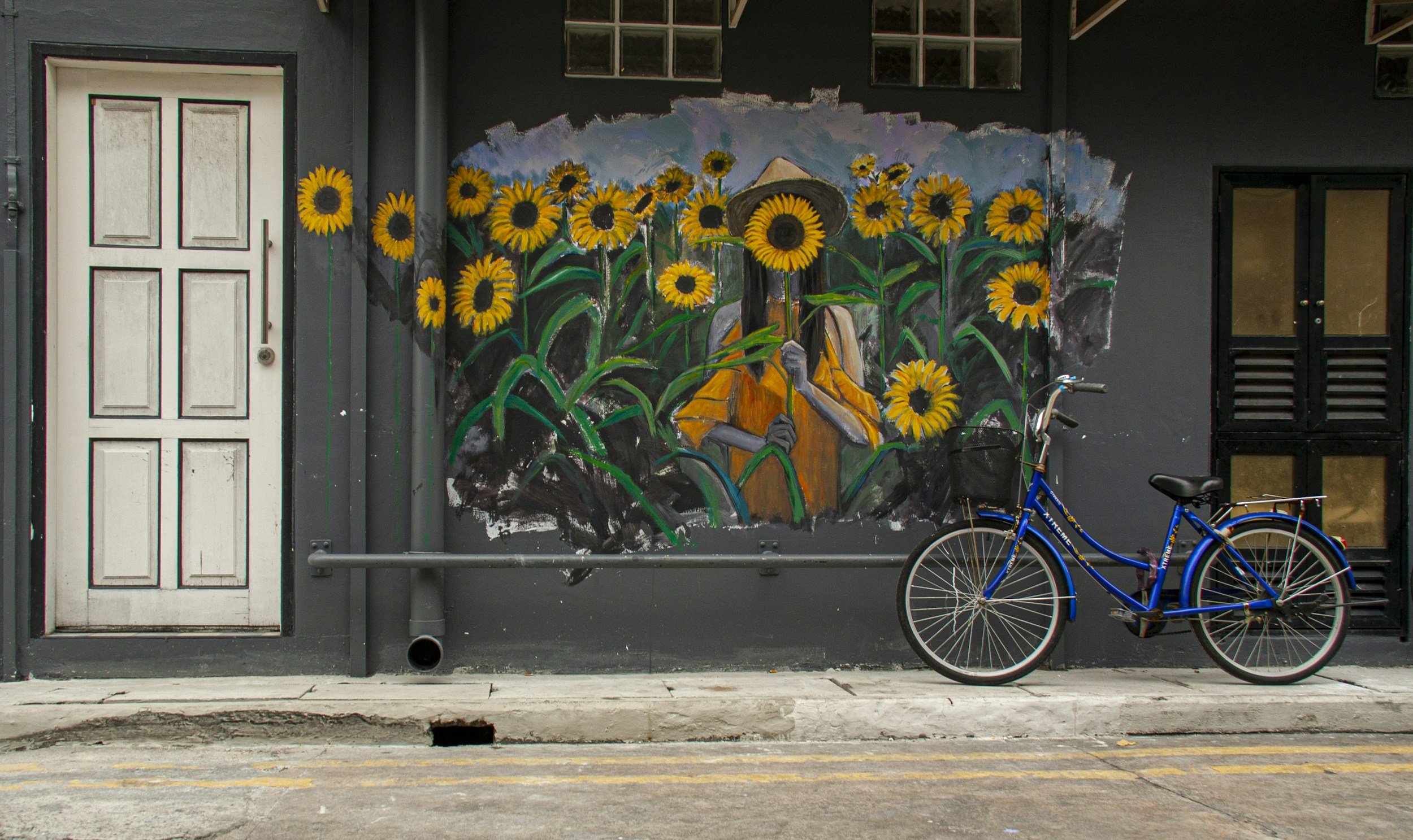 A mural painting of a person with a sunflower head, wearing a hat and yellow shirt, surrounded by sunflowers, on a dark background. There is a white door to the left and a black window with multiple panes to the right. A blue bicycle is parked in front.