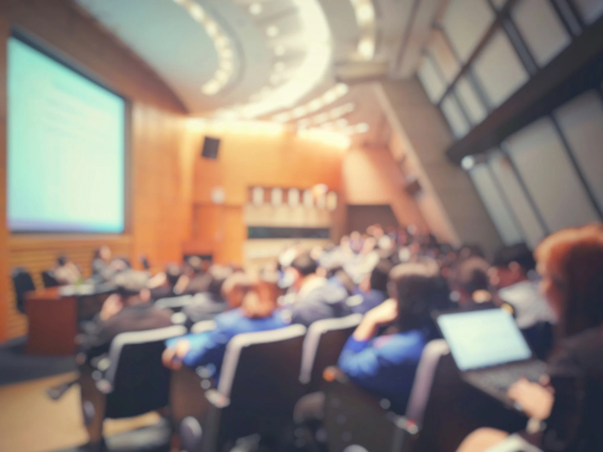 A large conference room filled with people sitting in rows, facing a stage with a large screen on the left, wooden walls, and a high ceiling with circular light fixtures.