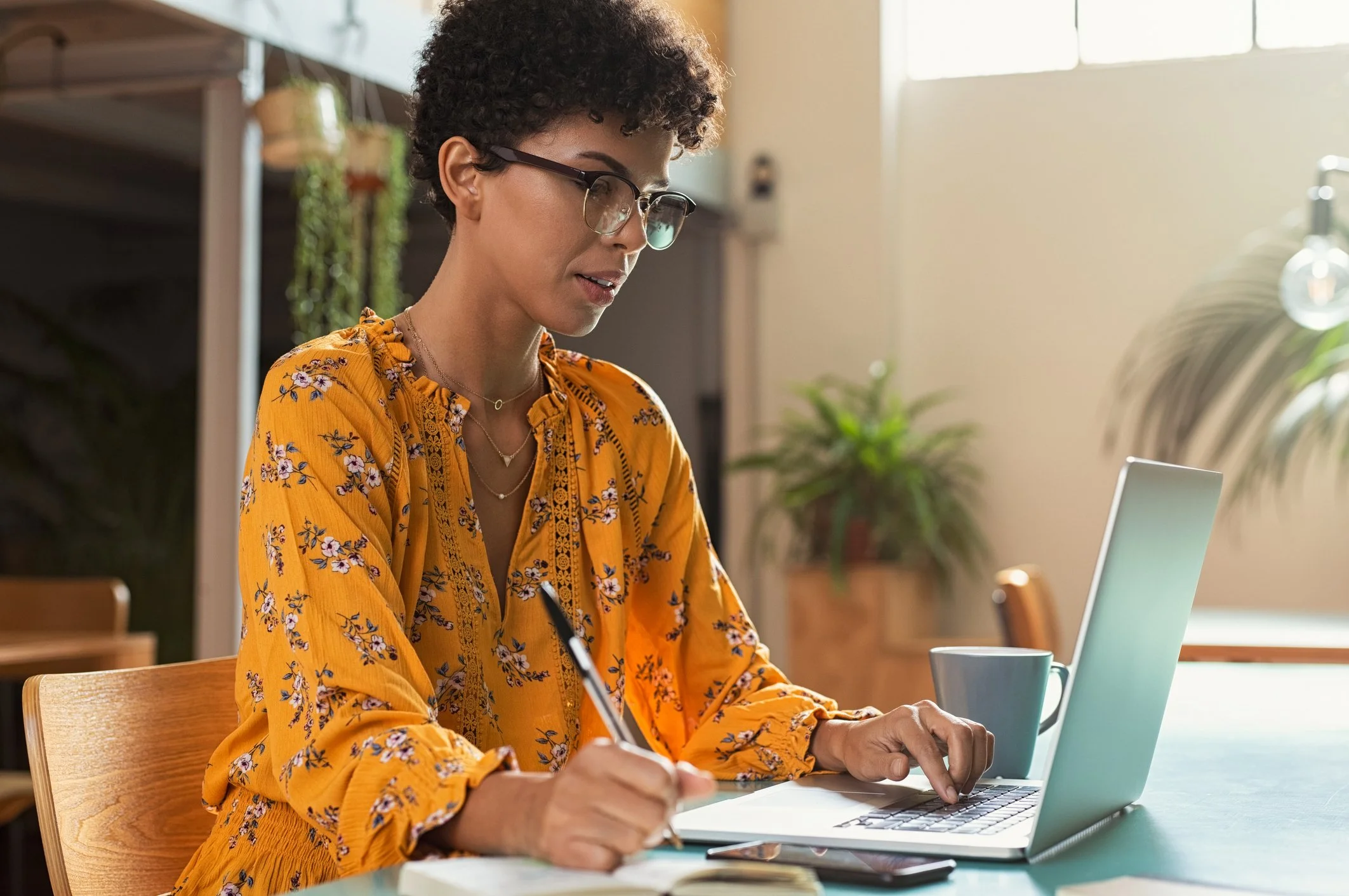 A woman with short curly hair, glasses, and a yellow floral blouse sitting at a table working on a laptop, with a notebook and pen in her hand, in a well-lit room with plants.