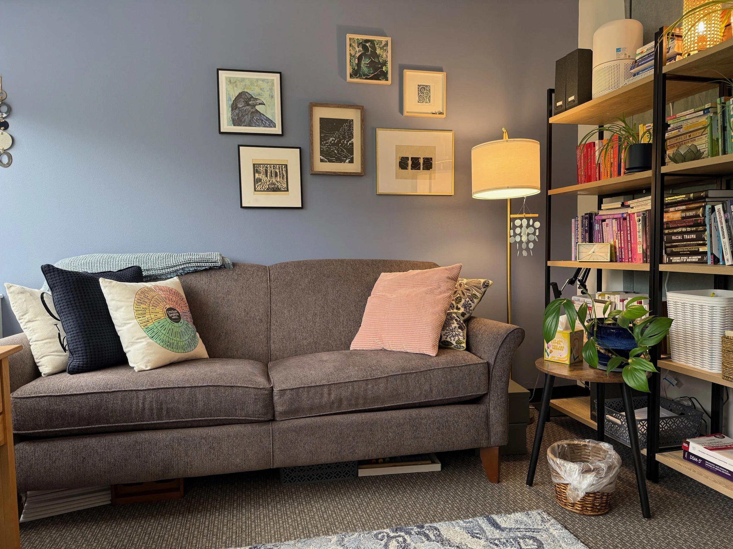 Photo of Laurie's therapy office in Mountlake Terrace WA with neutral large couch with pillows, neurodivergent-friendly floor lamp, plants, books, and framed art of fat bodies and other prints on the wall.