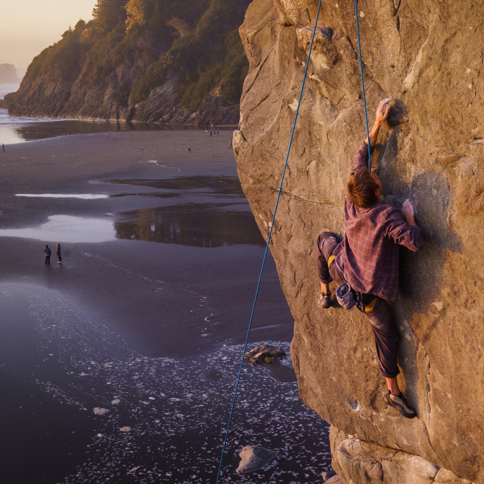 Kyle Fisher top-rope climbing a rock on the beach with puddles of sea water and trees in the background.