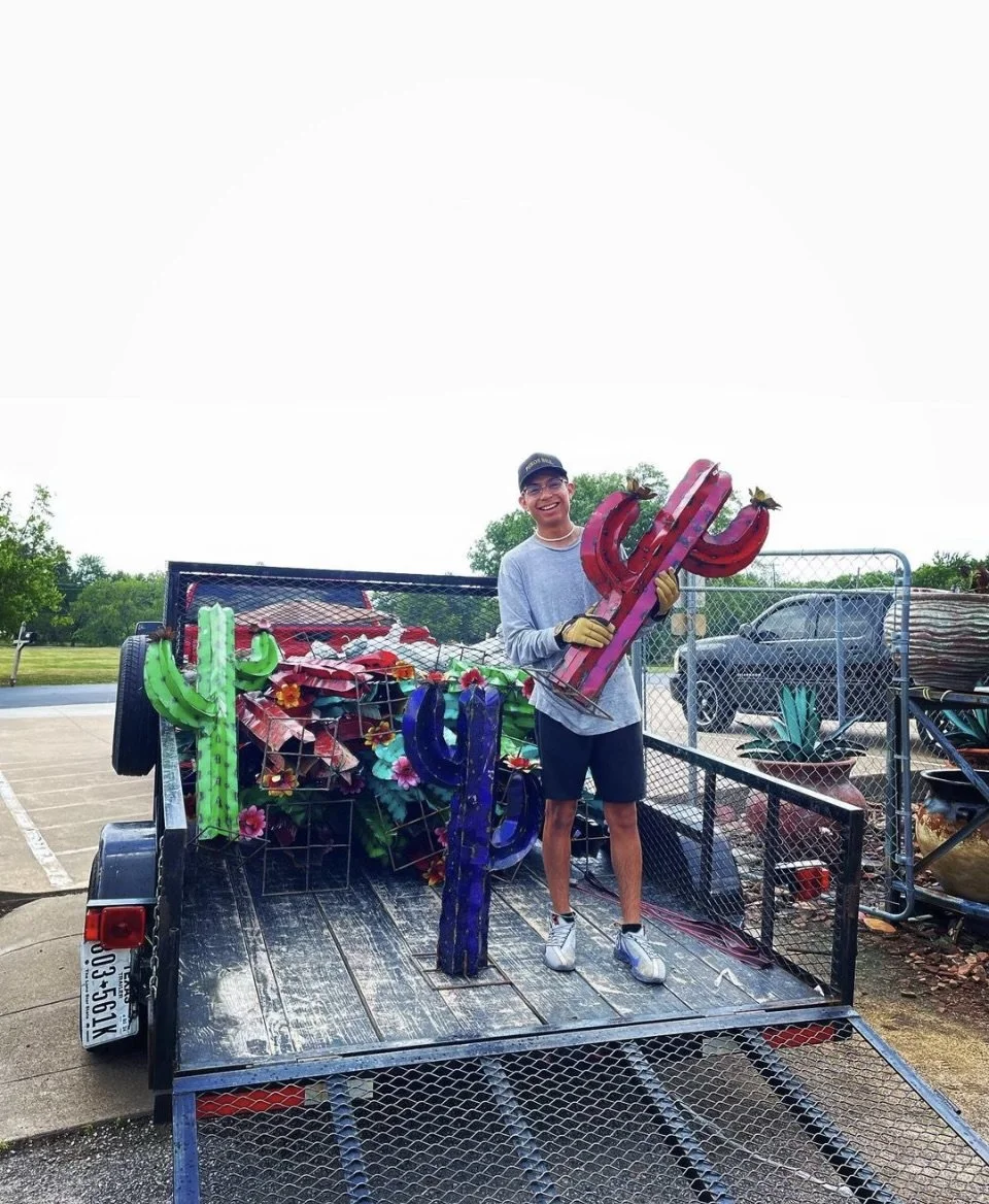 A young man in casual clothes and sunglasses standing on a trailer, holding a large pink and purple metal cactus sculpture, with bright green, blue, and purple similar sculptures attached to the trailer, outdoors on a cloudy day.