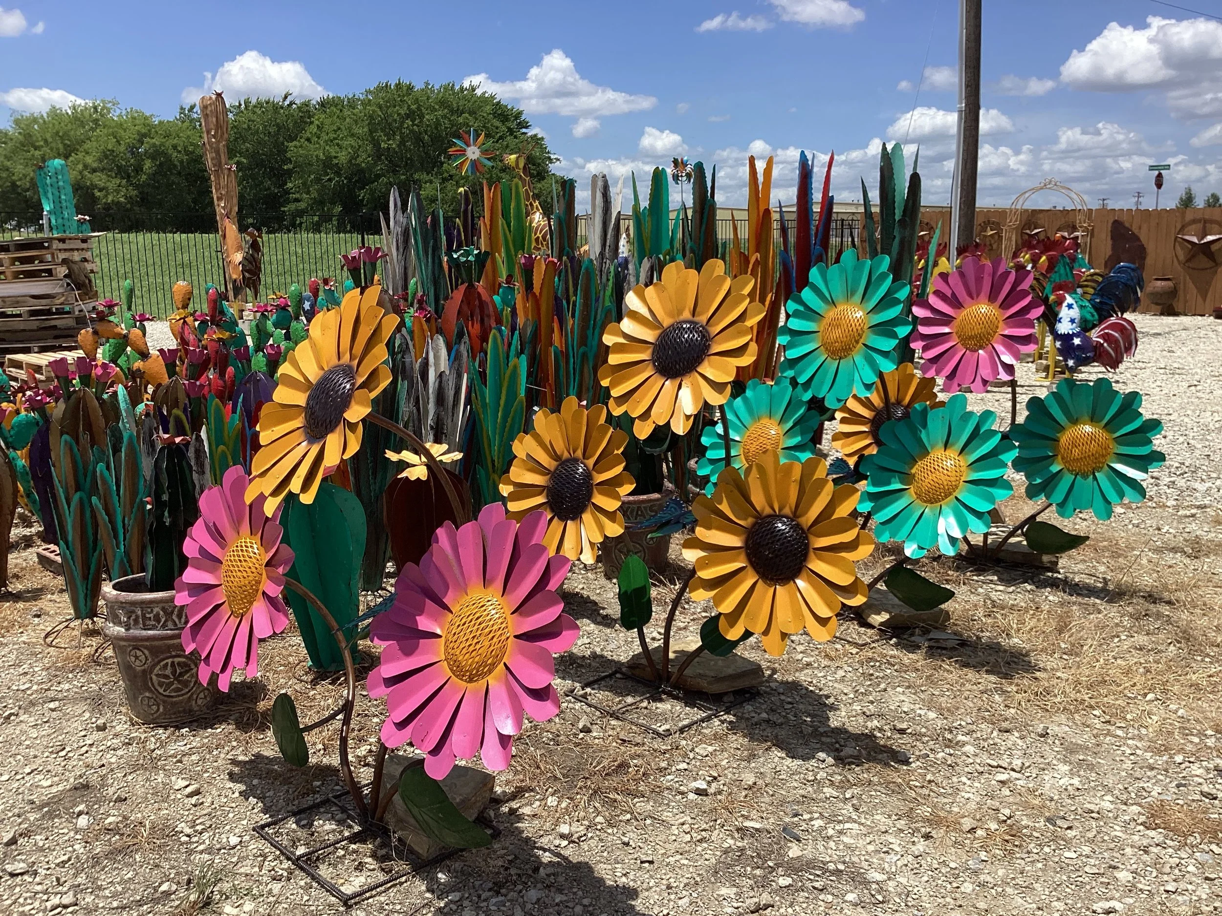 Colorful metal garden stakes in the shapes of pink, yellow, and teal flowers with black center, placed in gravel outdoors under a partly cloudy sky.