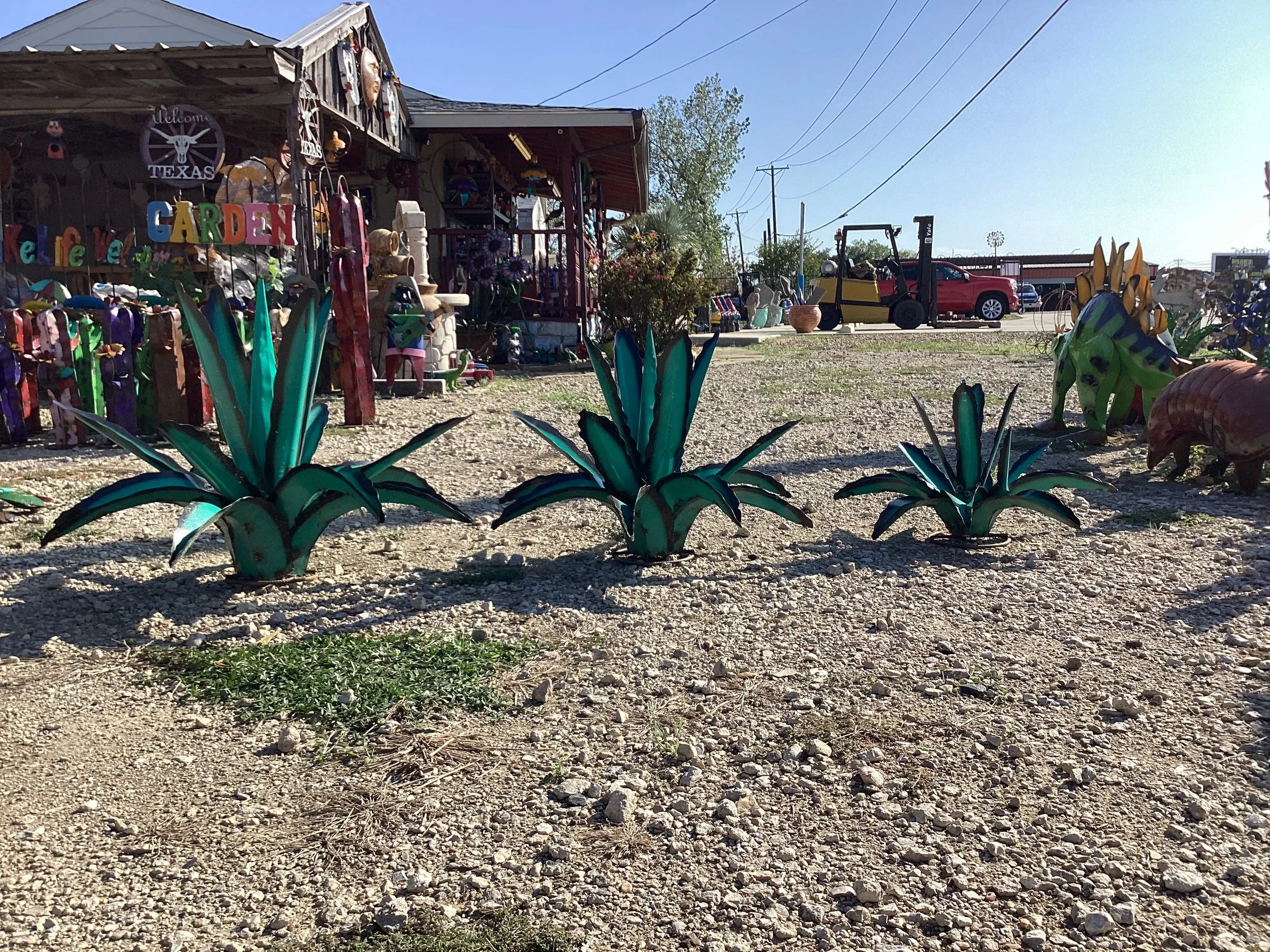 Colorful metal garden decorations resembling agave plants in front of a rustic shop with signs and other garden ornaments, on a gravel lot under a clear sky.