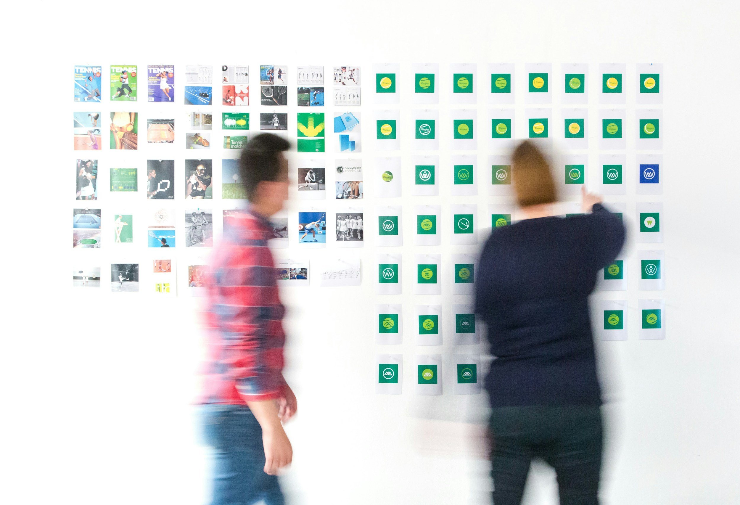 Two people in motion stand in front of a wall with various printed images and logos displayed in a grid pattern.