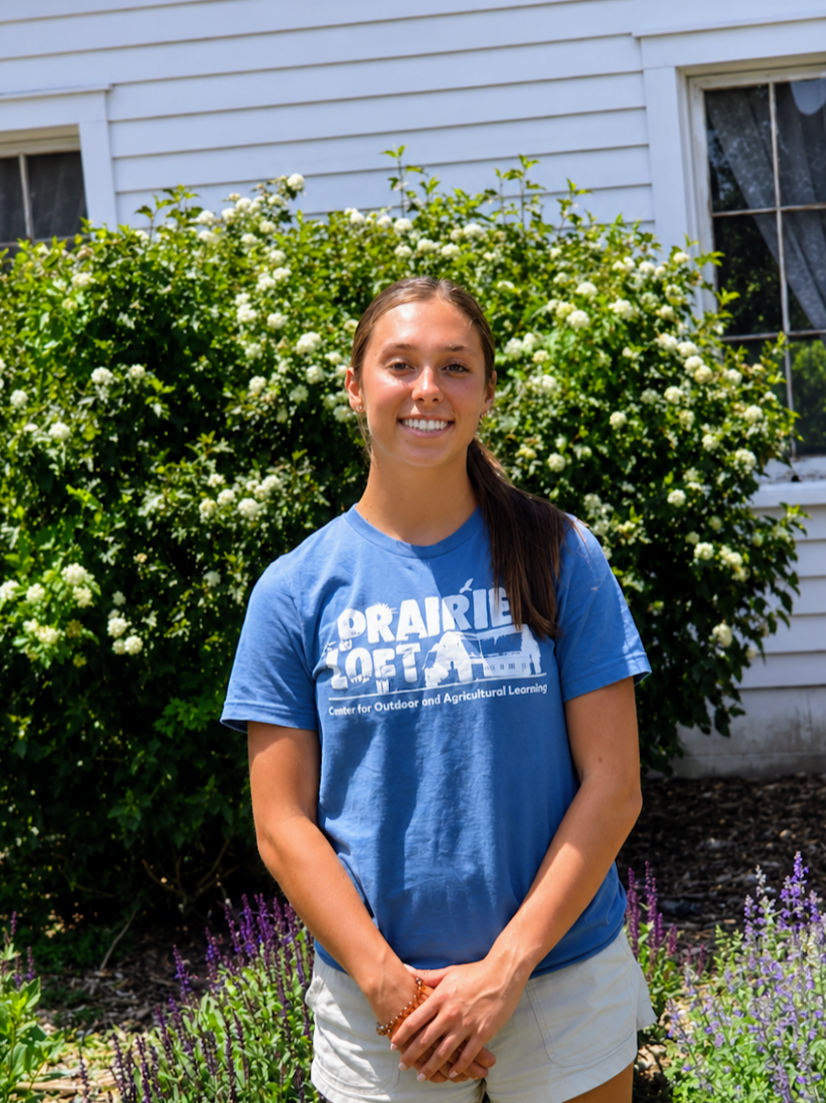 A young woman standing outdoors near a tree in a grassy area, wearing glasses, a pink t-shirt with the words 'Prairie Love' and a graphic of a prairie landscape, and blue jeans with a black belt. She is smiling at the camera.