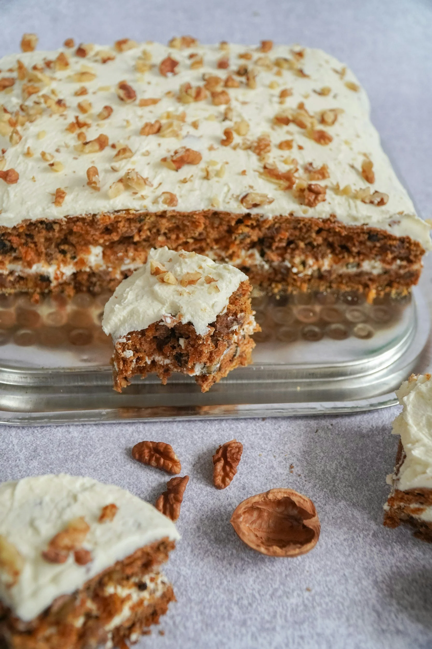 Carrot cake with cream cheese frosting topped with chopped walnuts, with slices and walnuts on the surface.