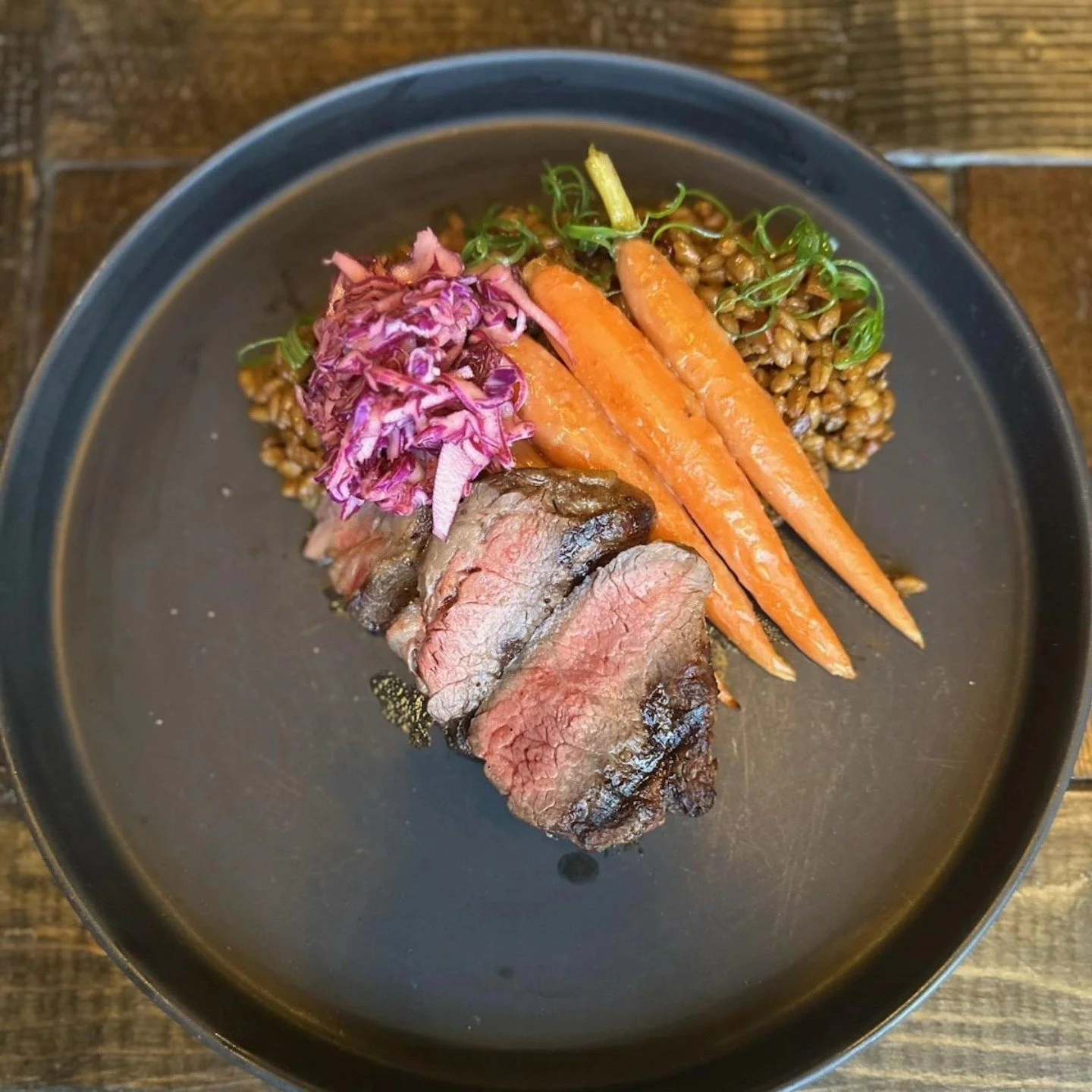 Slices of cooked beef, three whole carrots, purple cabbage slaw, cooked buckwheat, and microgreens on a black plate.