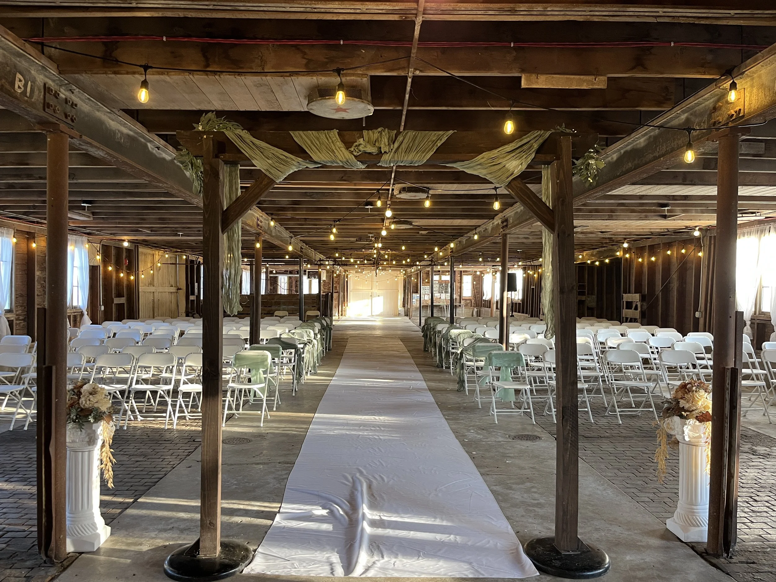 An indoor wedding ceremony setup with rows of white chairs, floral arrangements in glass vases, twinkle string lights, and wooden walls with windows, creating a rustic and romantic atmosphere.