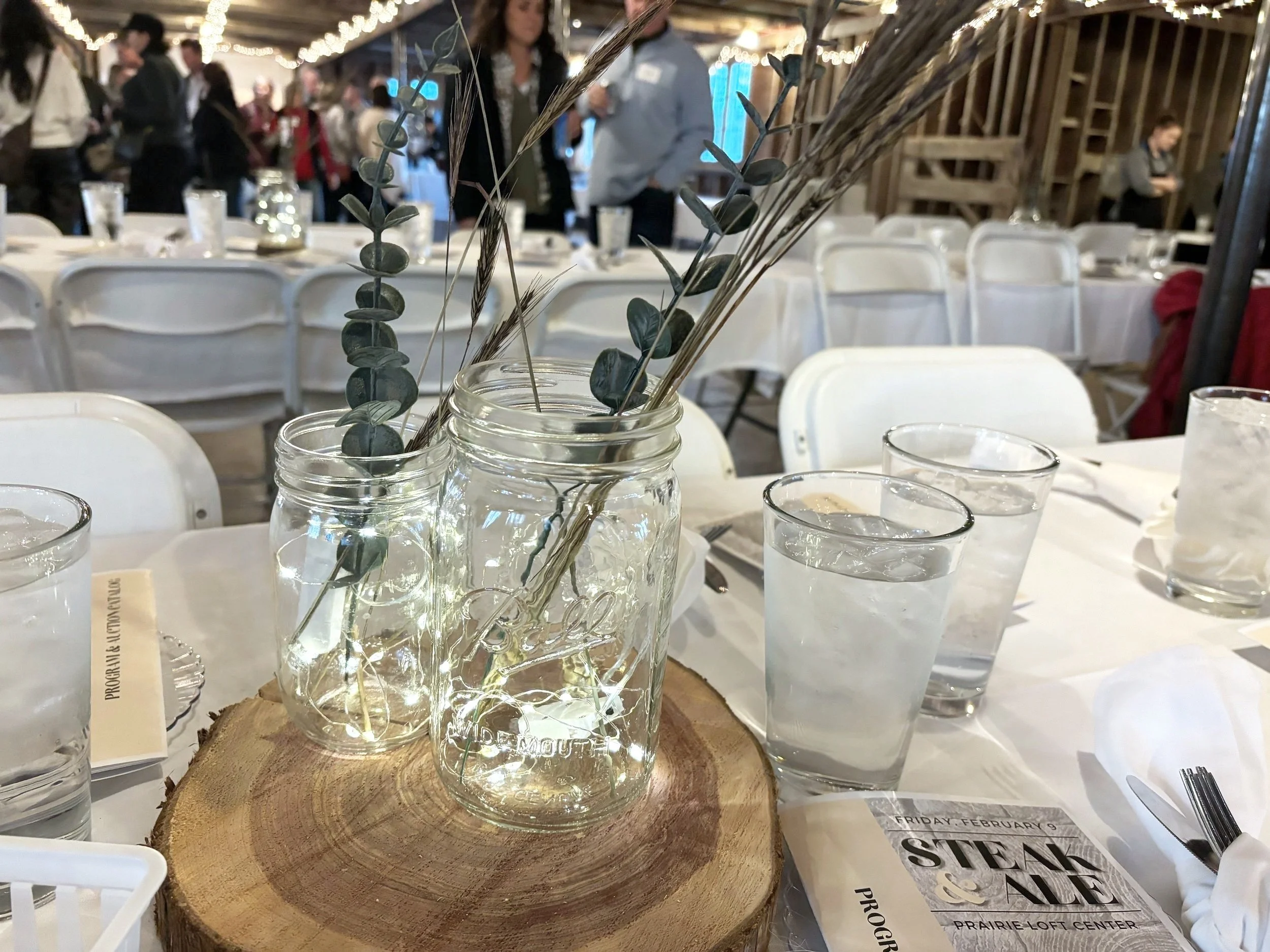 Table centerpiece with three glass jars containing eucalyptus leaves and dried grasses, placed on a wooden slab, surrounded by drinking glasses and tableware at a formal event.