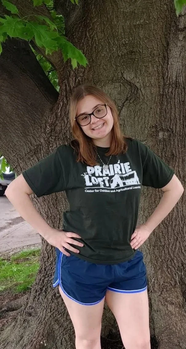 A young woman with long brown hair, smiling, wearing a green T-shirt with 'PRAIRIE LOFT' written on it, denim shorts, and a smartwatch, sitting in front of a red wooden wall.