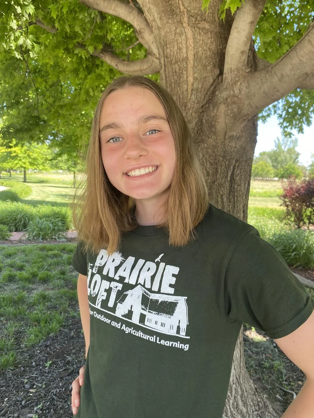 A young woman with long blonde hair smiling outdoors, wearing a blue t-shirt with the text 'Prairie Loft' and a name tag, sitting near greenery and buildings.