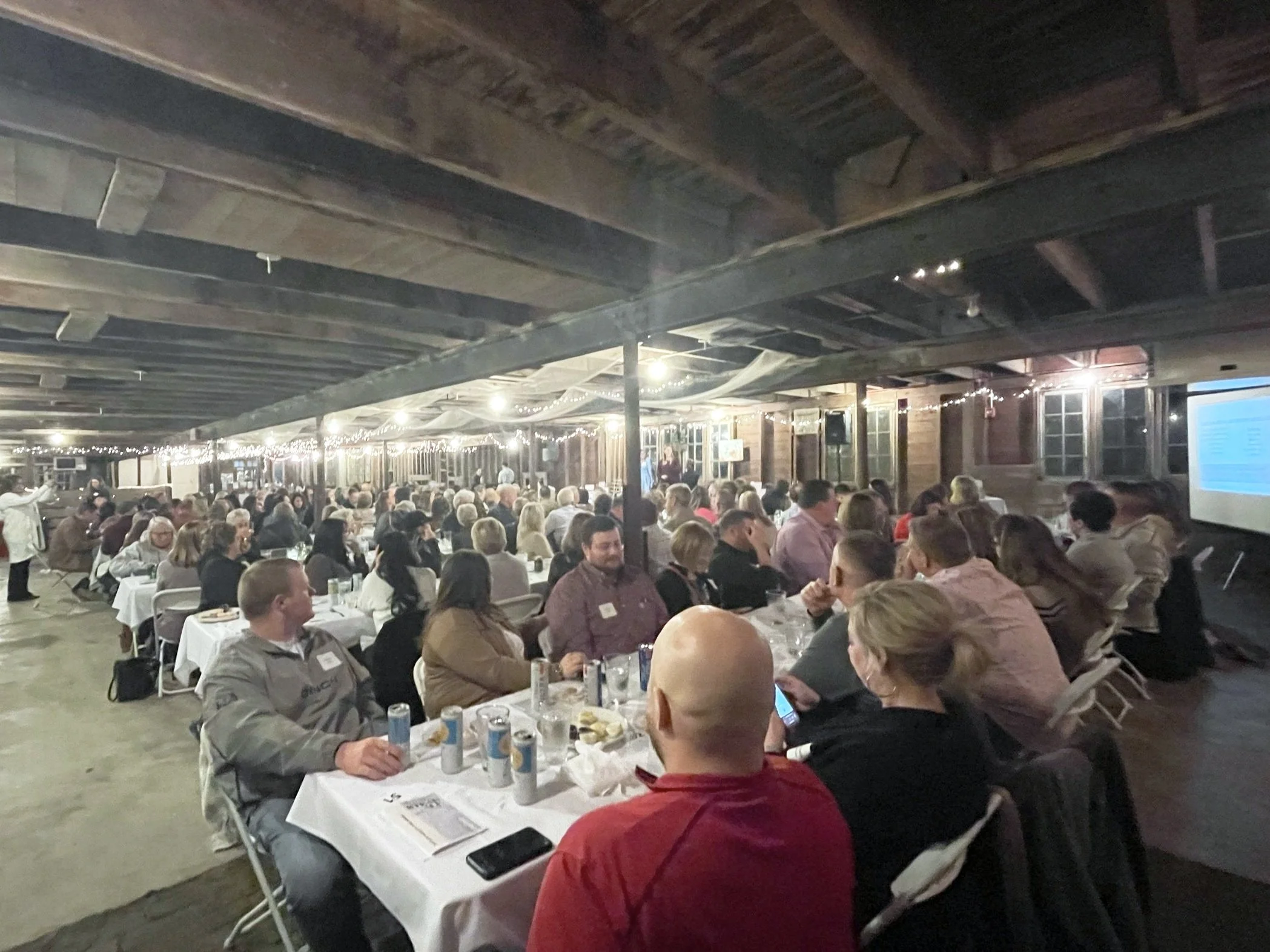People sitting at long tables with white tablecloths, dining and socializing in a rustic, barn-like venue decorated with string lights.