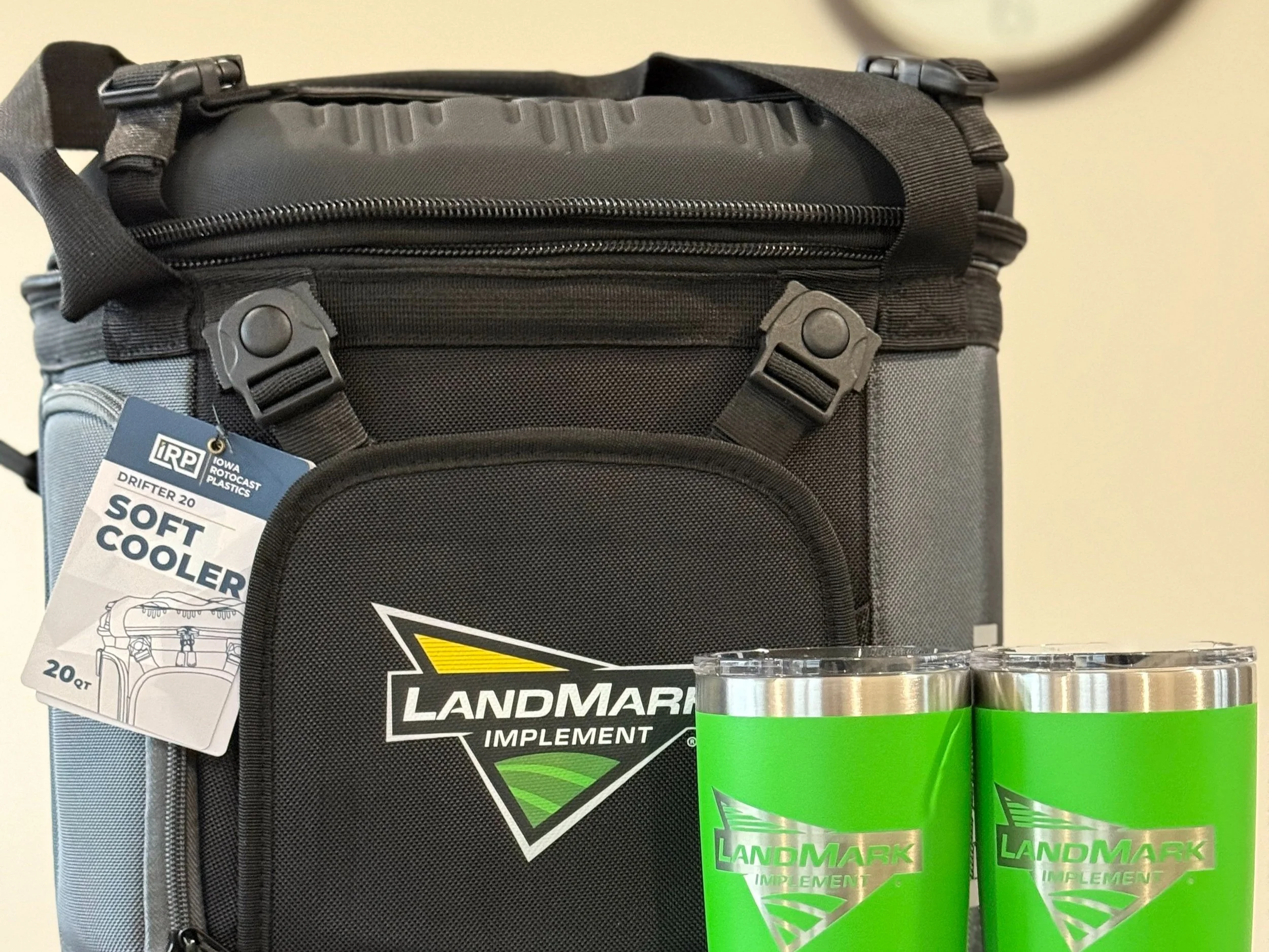 A black and gray cooler with Landmark Implement logo, two green cans of Landmark Implement branded soft cooler drinks, and a tag on the cooler reading 'DRIFTER 20 SOFT COOLER 20 QT'.