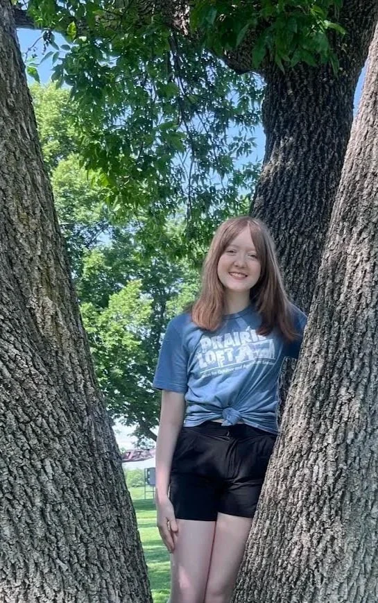 Prairie Loft Program Guides has long blonde hair standing in front of a lake and a tree, wearing a blue Prairie Loft t-shirt and denim shorts with floral patches.