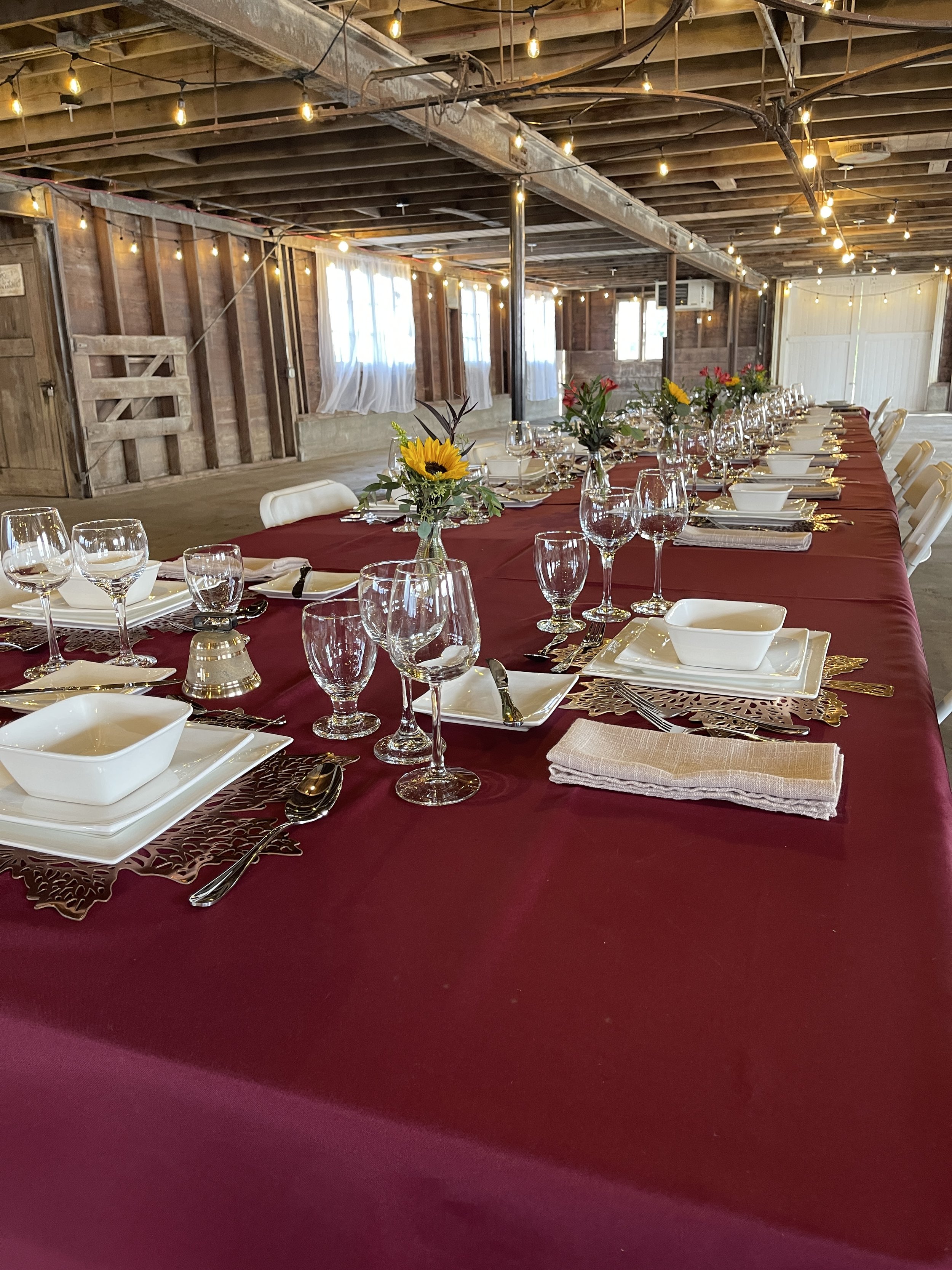 Long banquet table with a burgundy tablecloth, set with white plates, bowls, silverware, and various glasses. Centerpieces of sunflowers and other flowers decorate the table. The setting is in a rustic barn with wooden walls, string lights hanging from the ceiling, and natural light coming through windows.