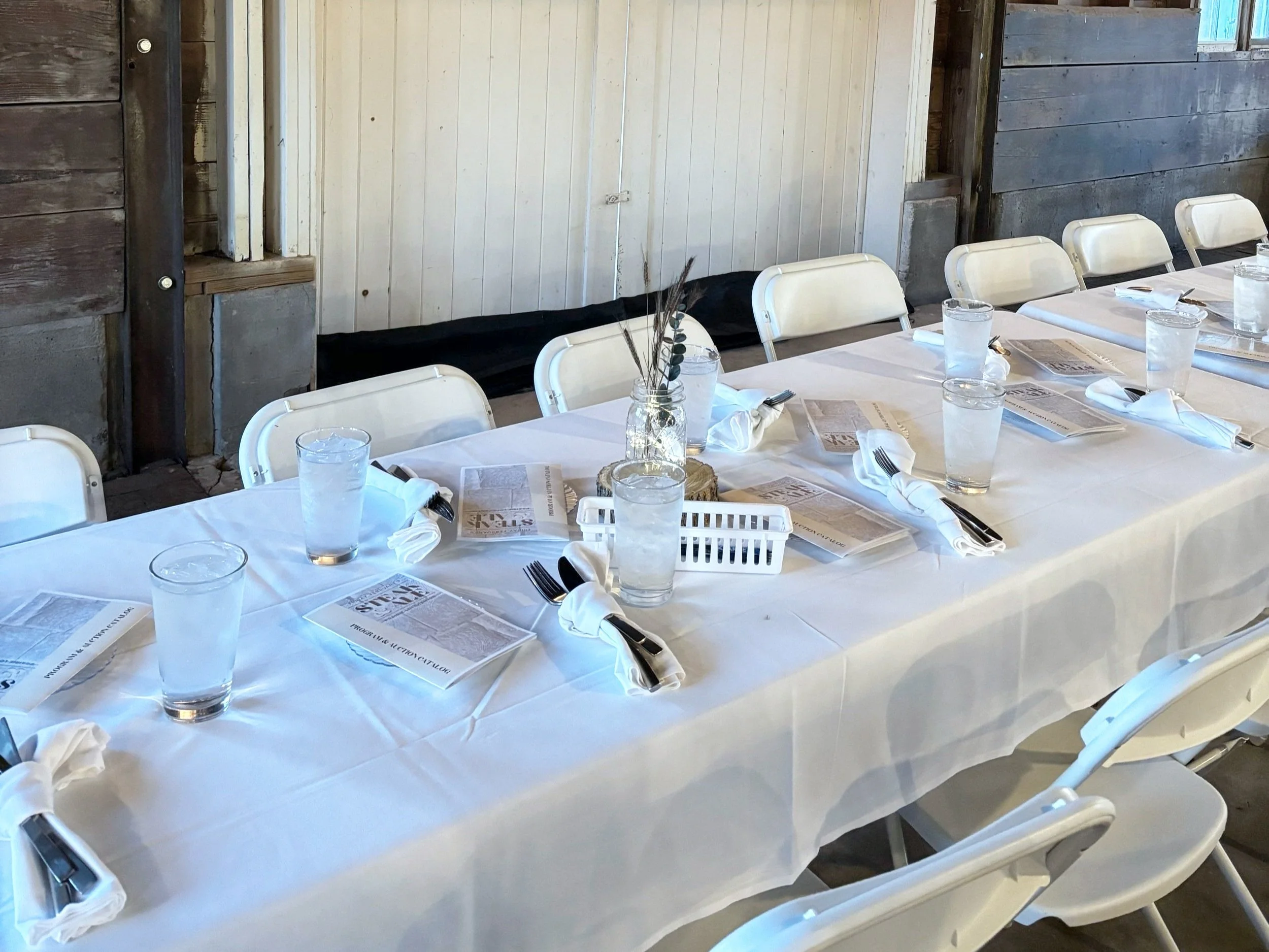 Table set for a meal with white tablecloth, water glasses, napkins, and menus, in a rustic wooden room.