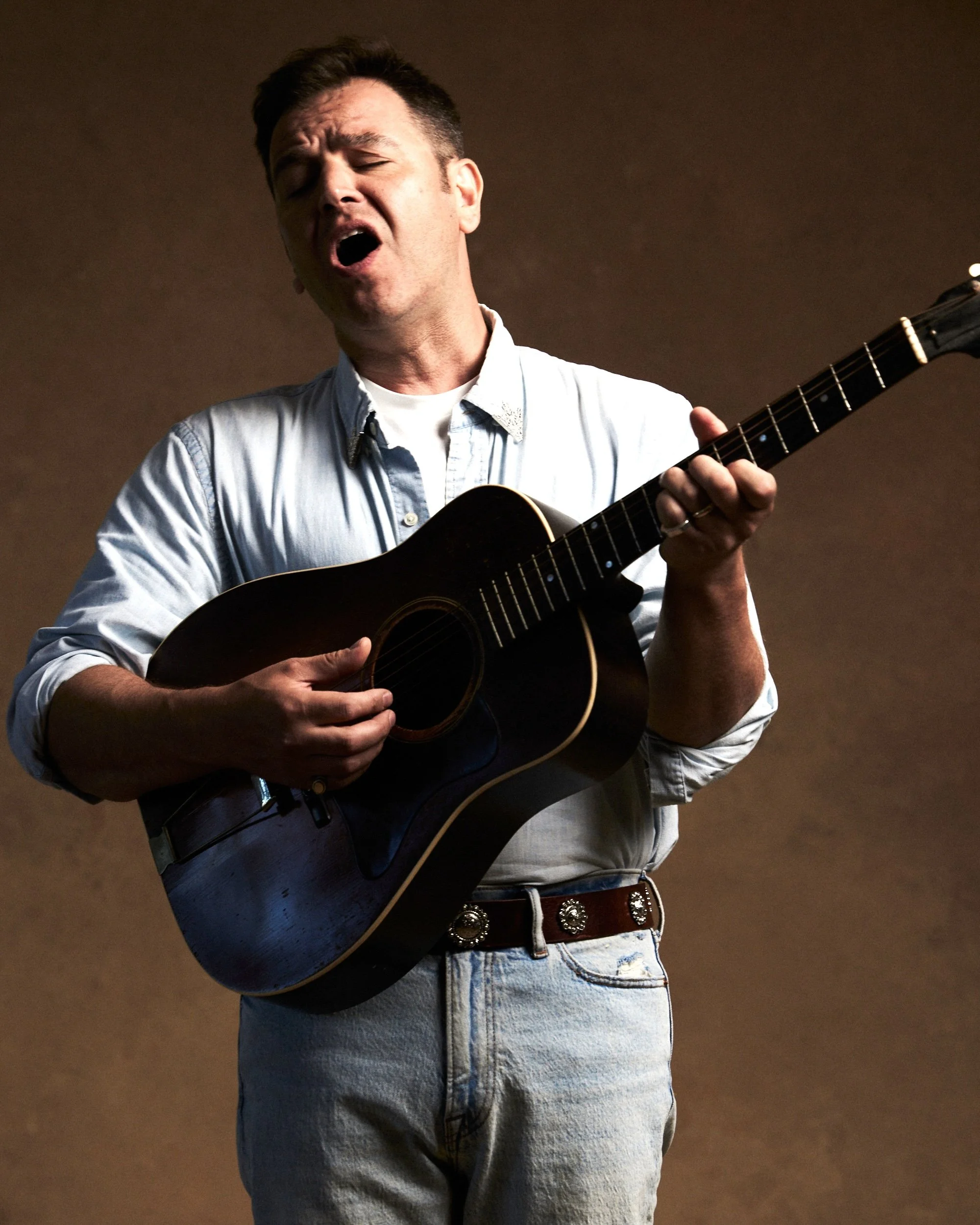 A man playing an acoustic guitar, singing, with a dark background. He's wearing a light denim shirt and light-colored jeans with a decorative belt.