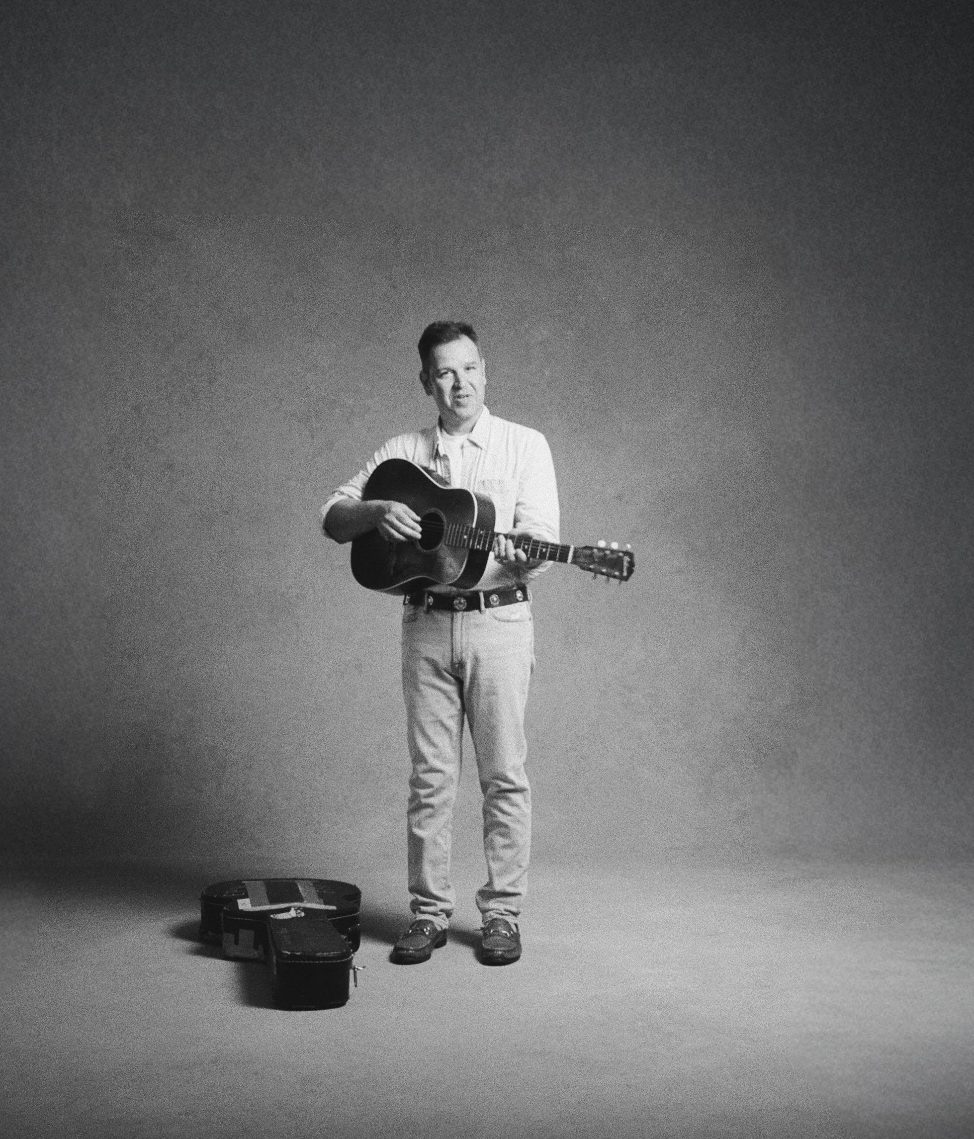 Black and white photo of a man playing an acoustic guitar, dressed in casual clothing, standing on a plain background with a guitar case on the floor nearby.