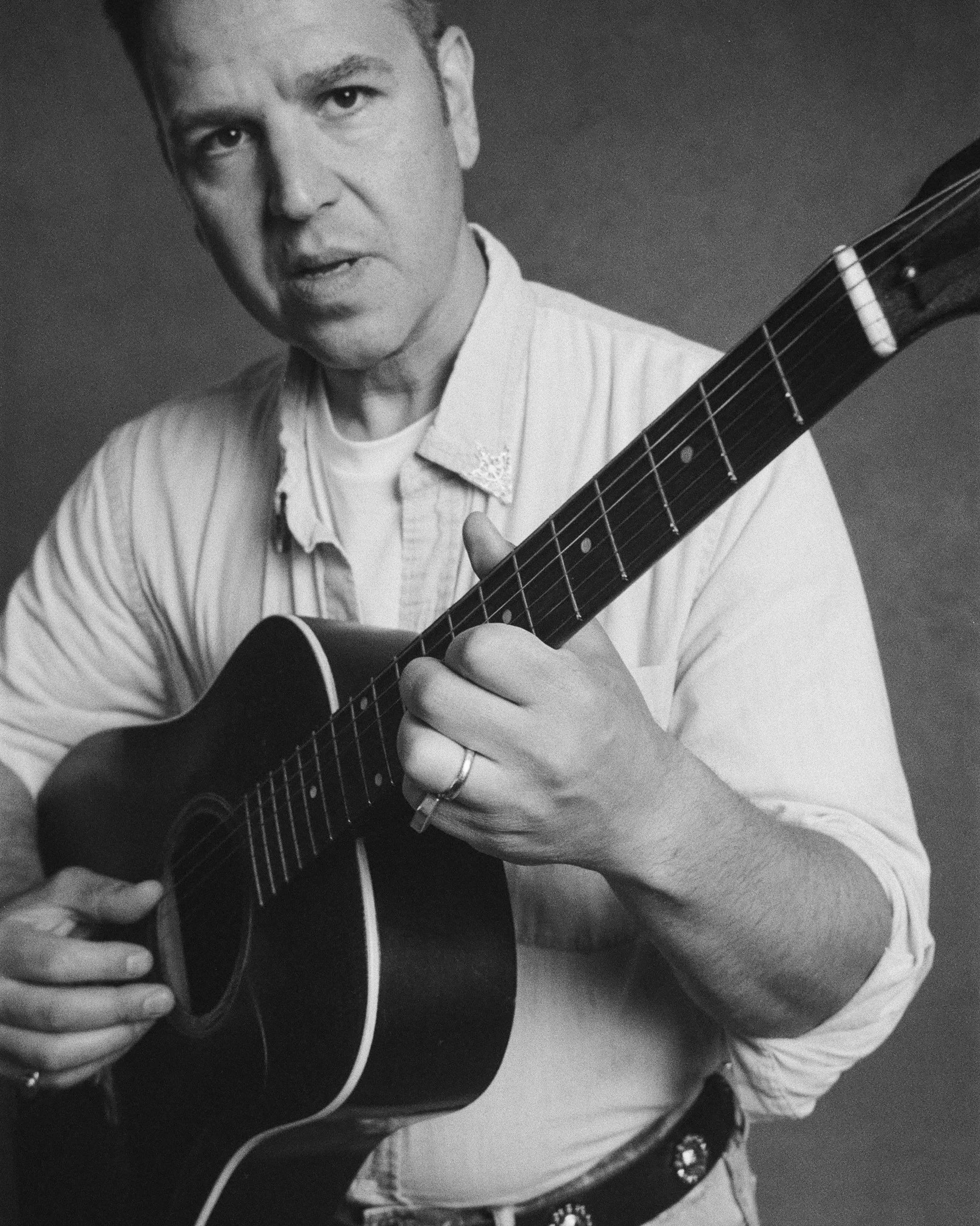 A black-and-white photo of a man playing an acoustic guitar, wearing a light-colored, striped shirt, with a ring on his finger and a belt with a decorative buckle, against a plain background.