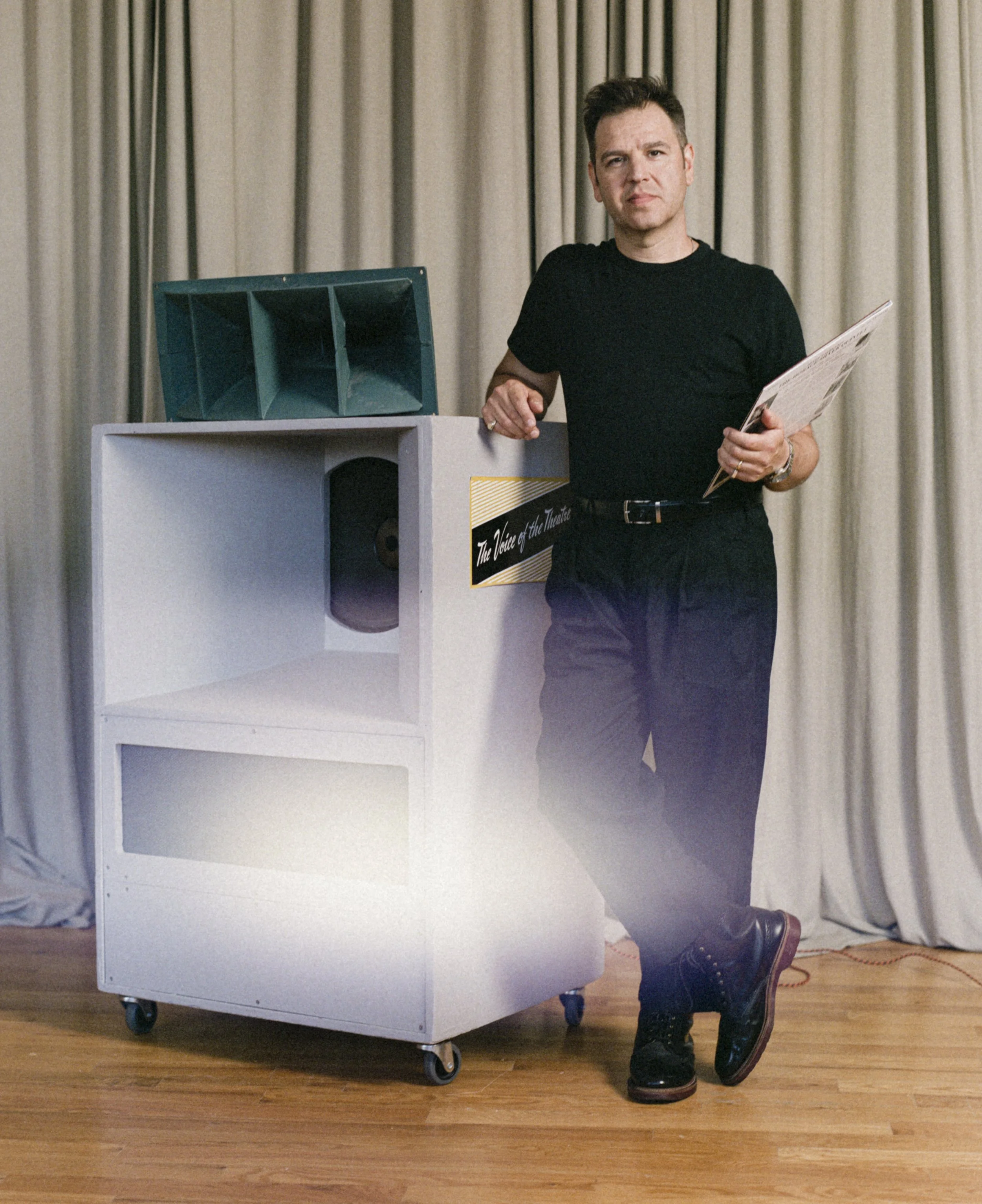 Person standing behind a white podcaster's booth labeled 'The Voice of the Theatre,' wearing black pants, black shoes, and a black T-shirt, holding papers, with curtains in the background and a wooden floor.