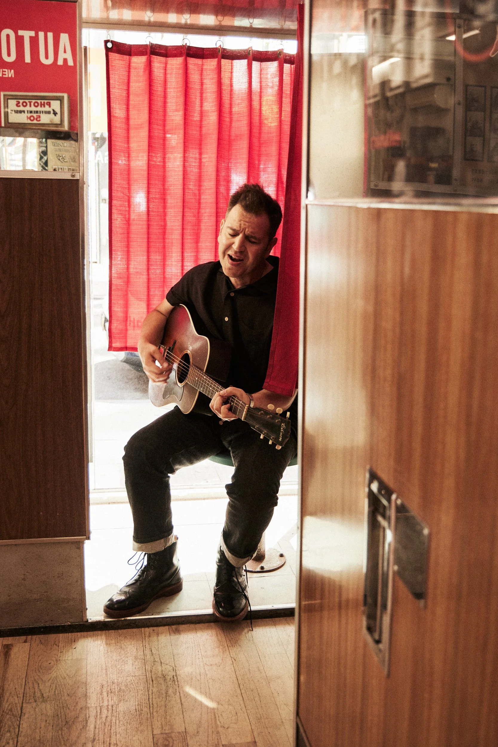 A man playing an acoustic guitar in a small, cozy restaurant or cafe, sitting in front of a red curtain with a window behind him.