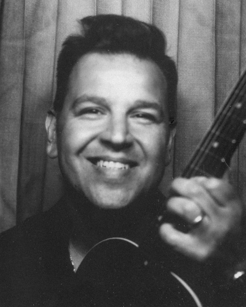 Black-and-white photo of a smiling man holding a guitar, standing in front of a wooden wall.