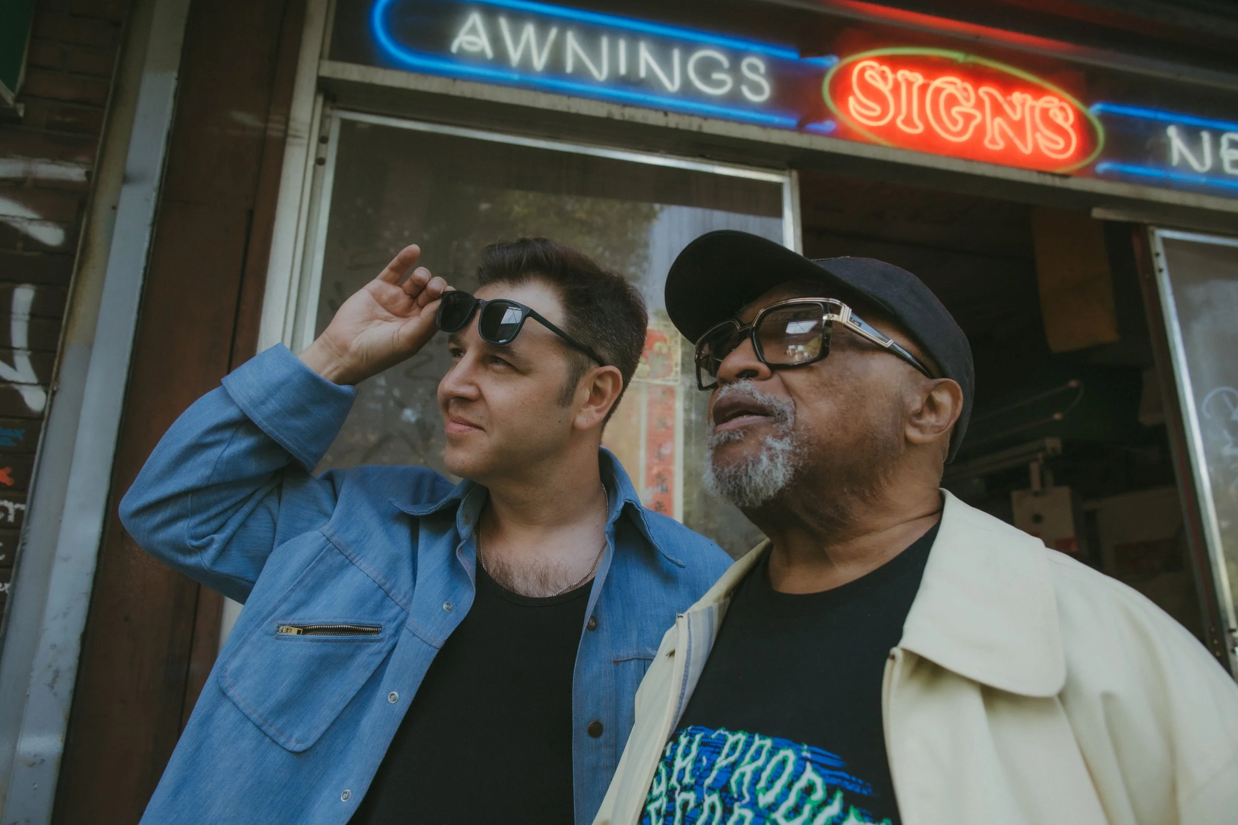 Two men standing outside a storefront with neon signs reading 'AWNINGS' and 'SIGNS.' The younger man on the left is wearing sunglasses, a blue denim shirt, and is adjusting his sunglasses. The older man on the right is wearing glasses, a cap, and a light-colored jacket with a colorful patterned shirt underneath.