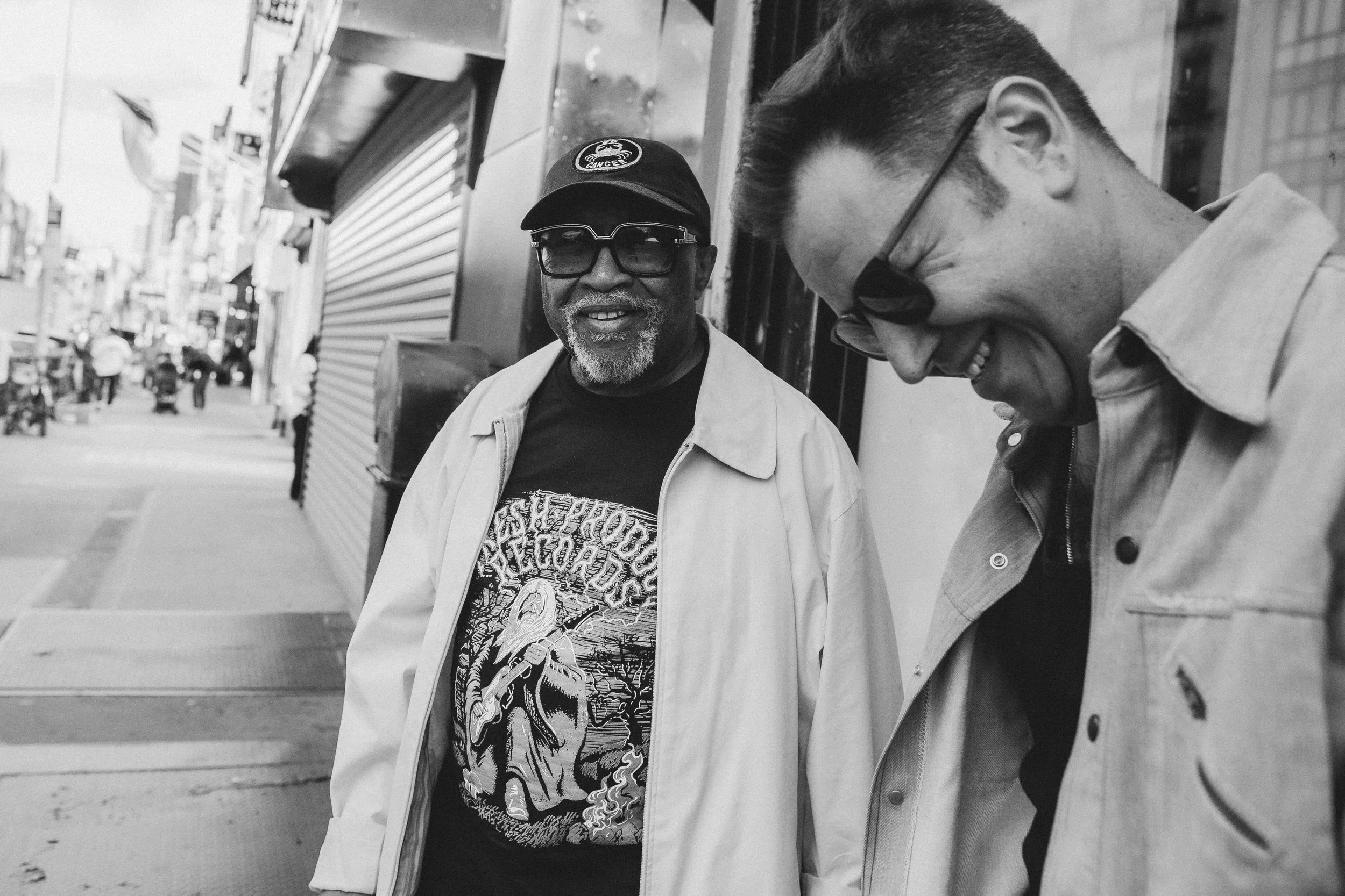 Two men standing and talking on a city street in black and white, one wearing glasses and a cap, the other with hair styled back and a collared shirt, with storefronts and pedestrians in the background.