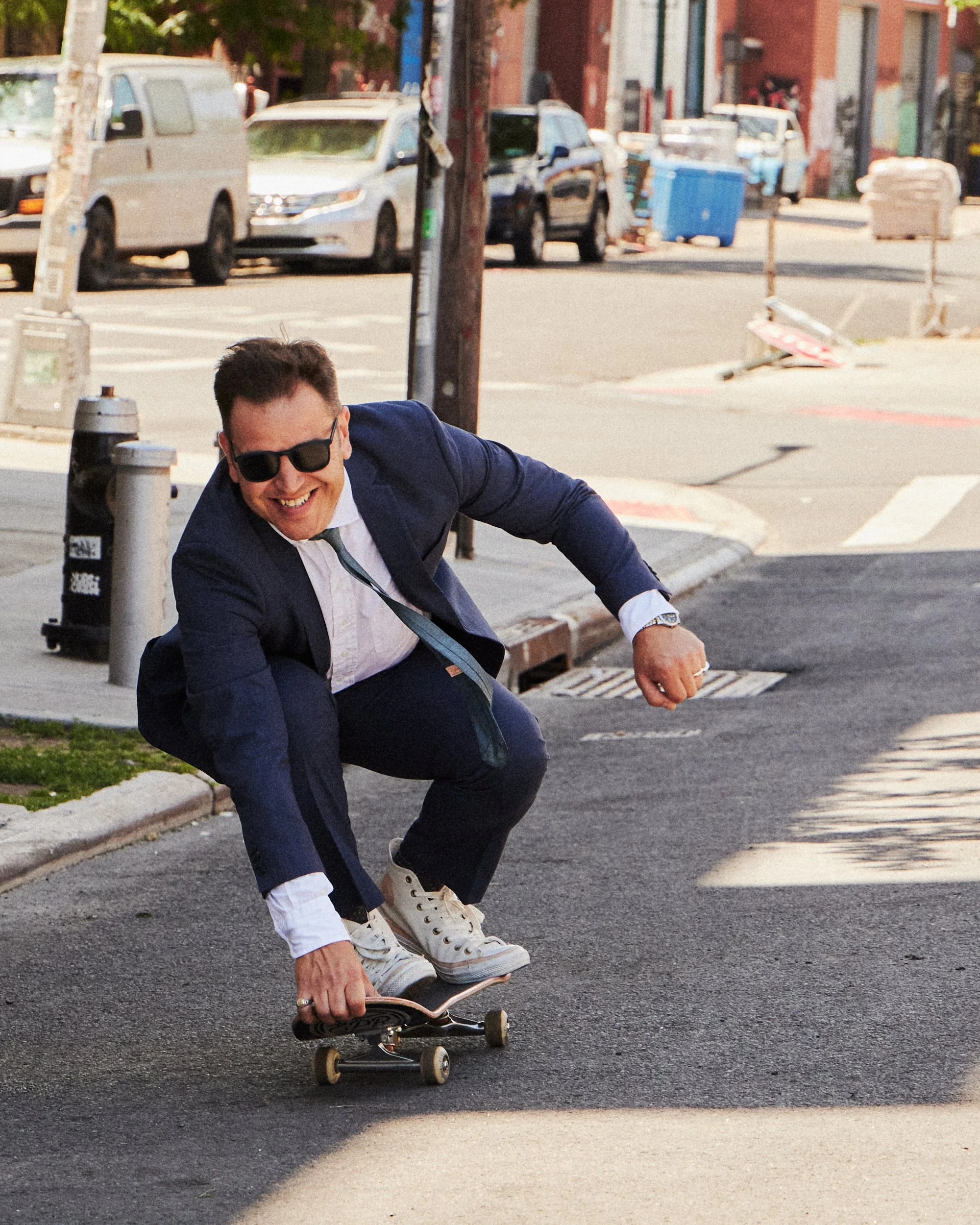 A man wearing a dark suit, white shirt, and sunglasses is crouching down with a skateboard on a city street, smiling.