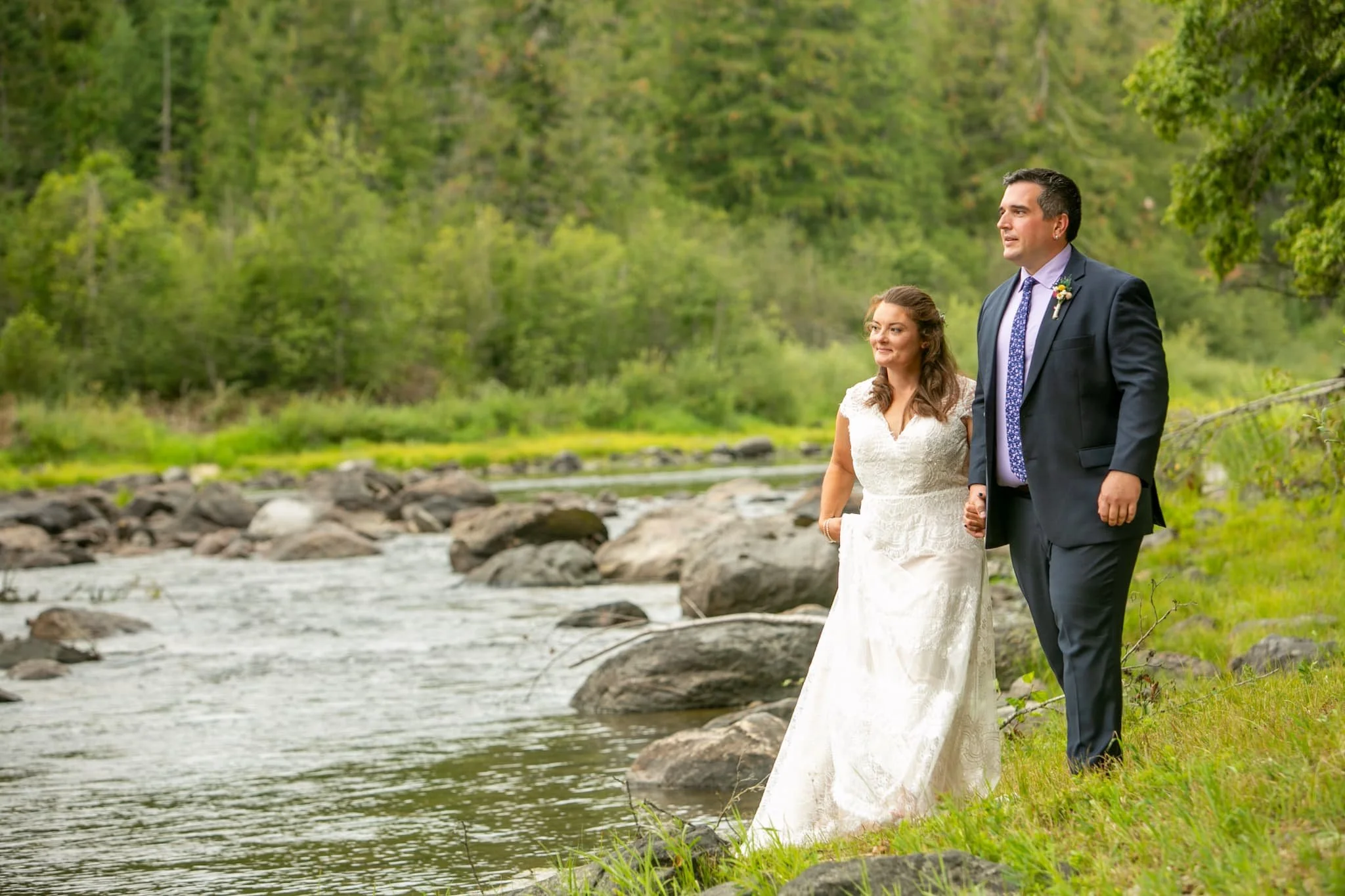 Bride and groom walking along the riverbank at our Northern Idaho venue, the forest and river creating a dramatic, natural backdrop for an edgy outdoor portrait.