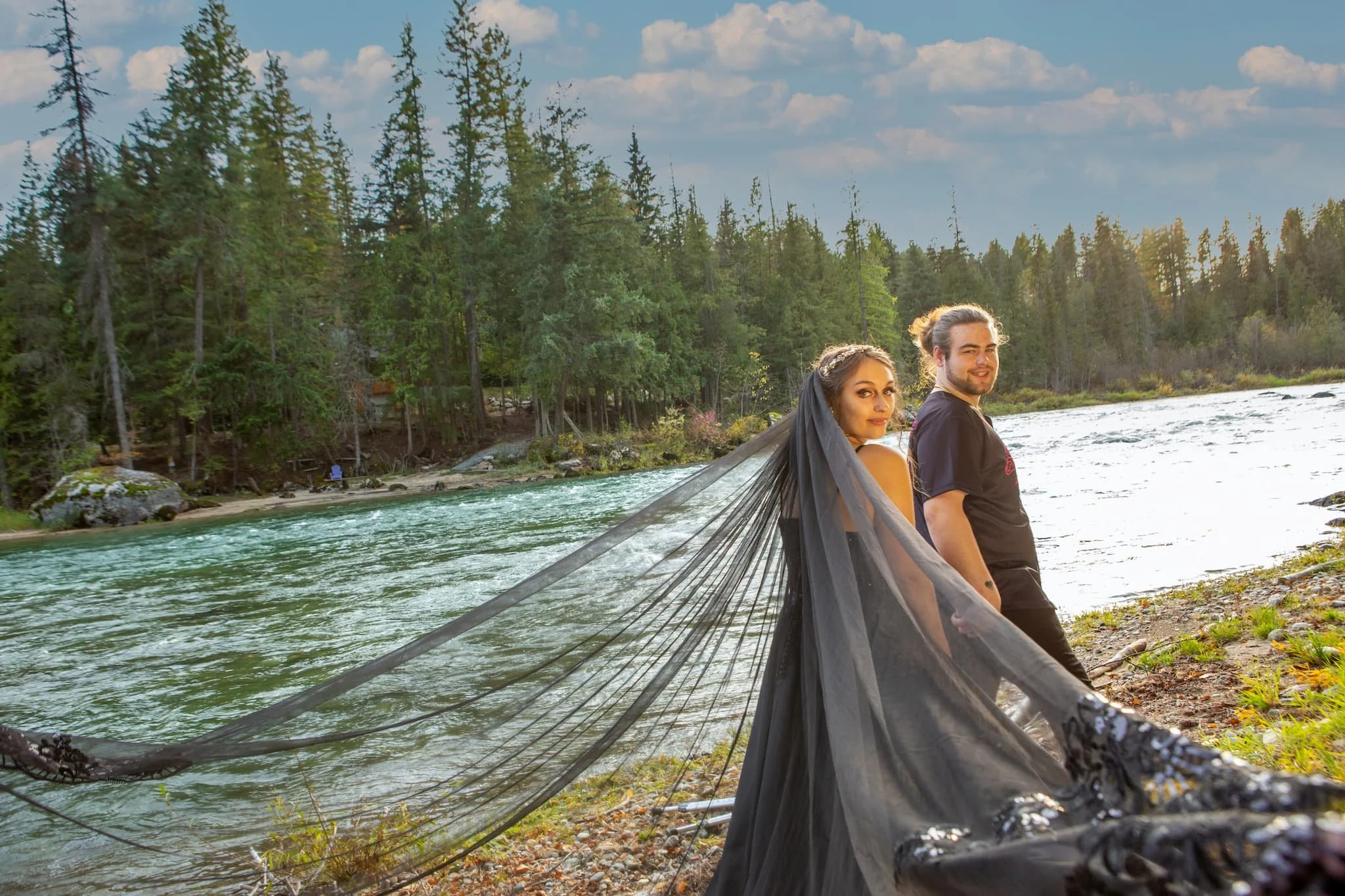 The couple walks along a waters edge at our Priest River riverfront venue, the soft light of golden hour and towering trees creating a moody, cinematic wedding moment.