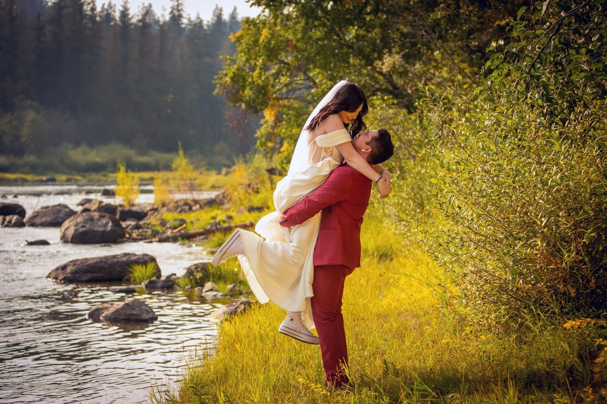 Couple sharing a romantic embrace along the river’s edge at Cedar River Wedding Venue in Priest River, Idaho, framed by towering cedar trees and soft golden light.