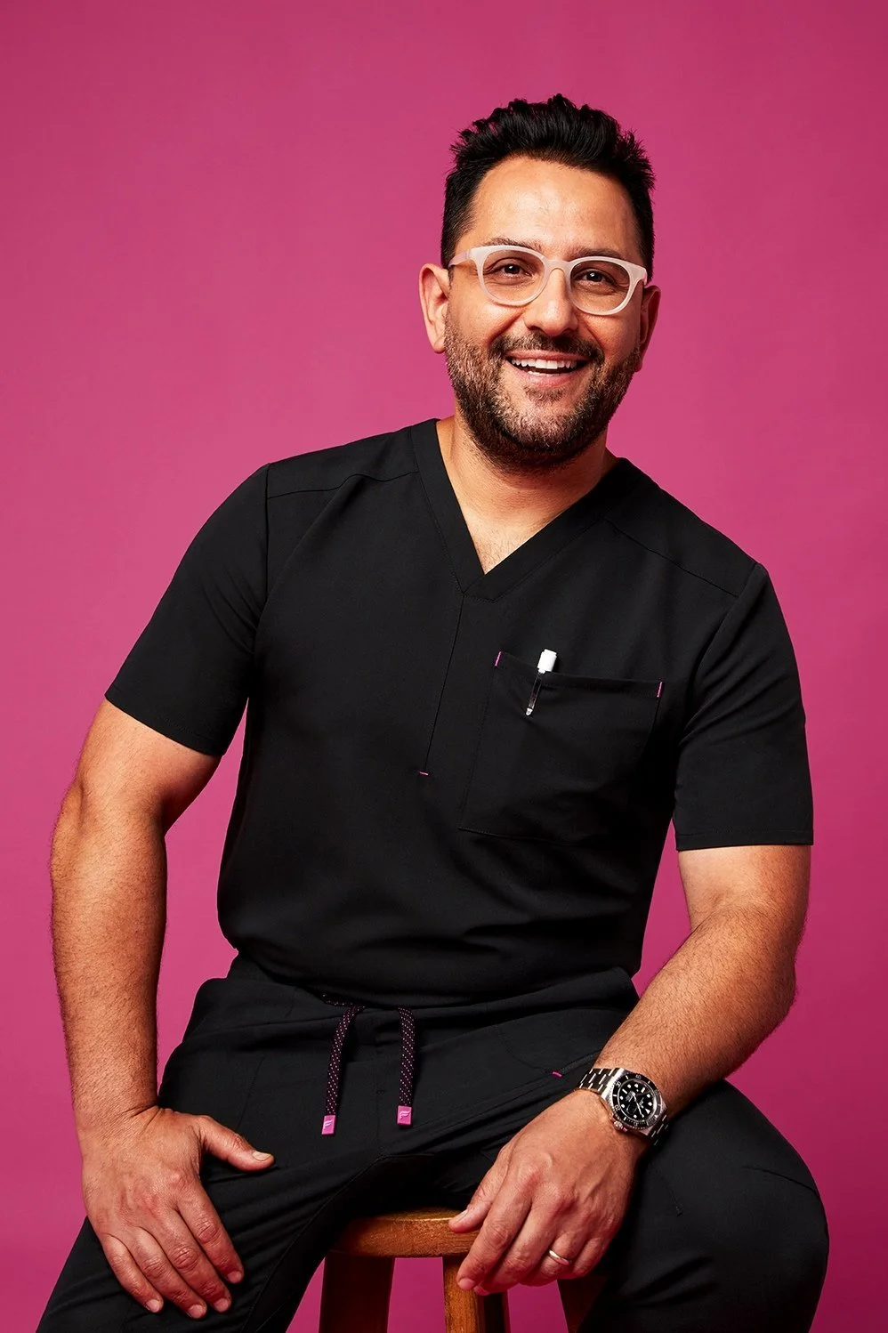 Man with dark hair and beard, wearing glasses and black medical scrubs, sitting on a wooden stool against a pink background, smiling at the camera.