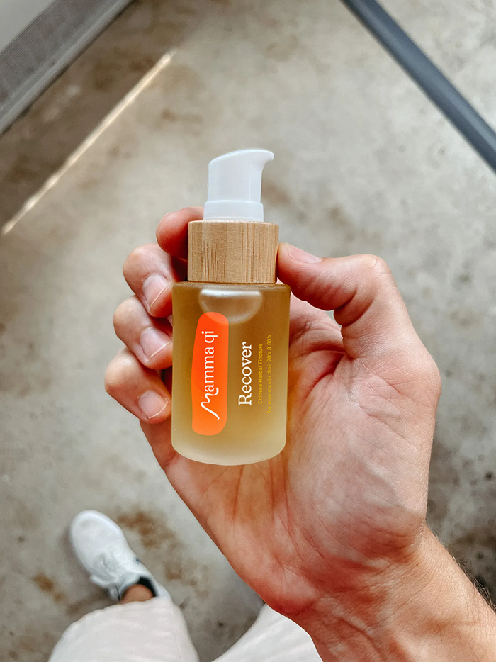 Hand holding a small bottle of skincare serum labeled 'Mama Gi Recovery' with a pump dispenser, against a background of concrete floor and part of a shoe.