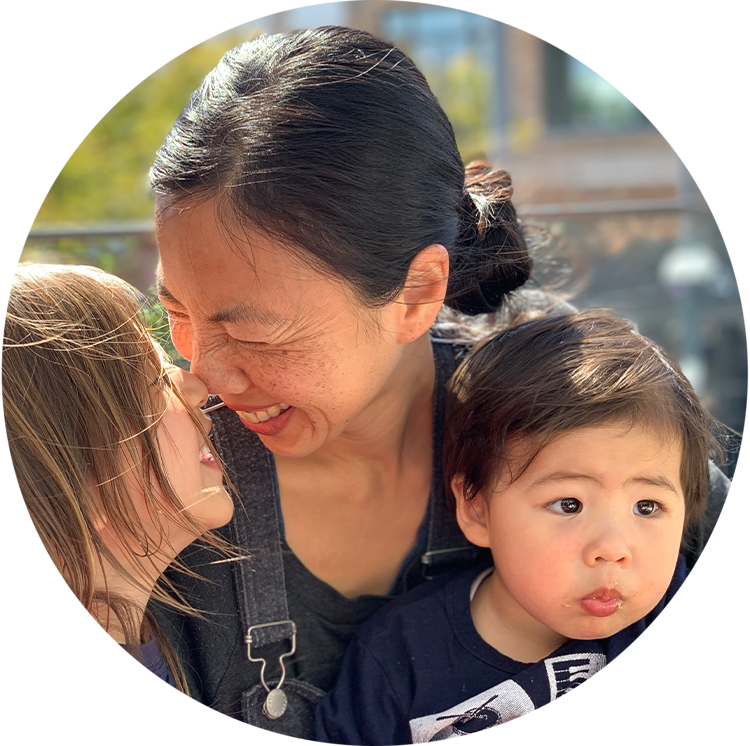 A woman with two children, one girl and one boy, outdoors on a sunny day. The woman and girl are smiling and touching noses, while the boy looks to the side with his lips pursed.