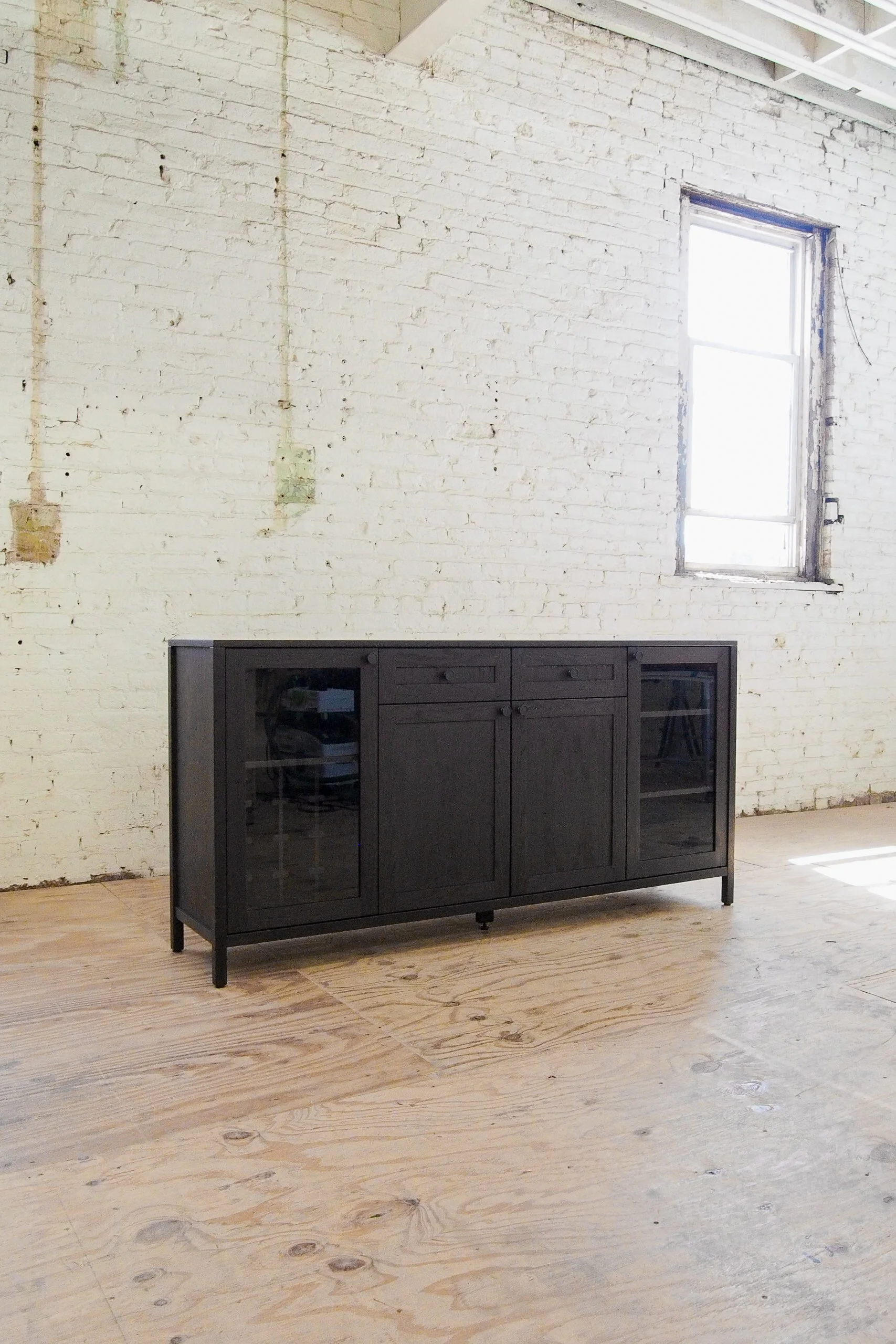 A black cabinet with glass doors and drawers is positioned against a white painted brick wall with an old window. The scene has wooden flooring and natural light streaming in.