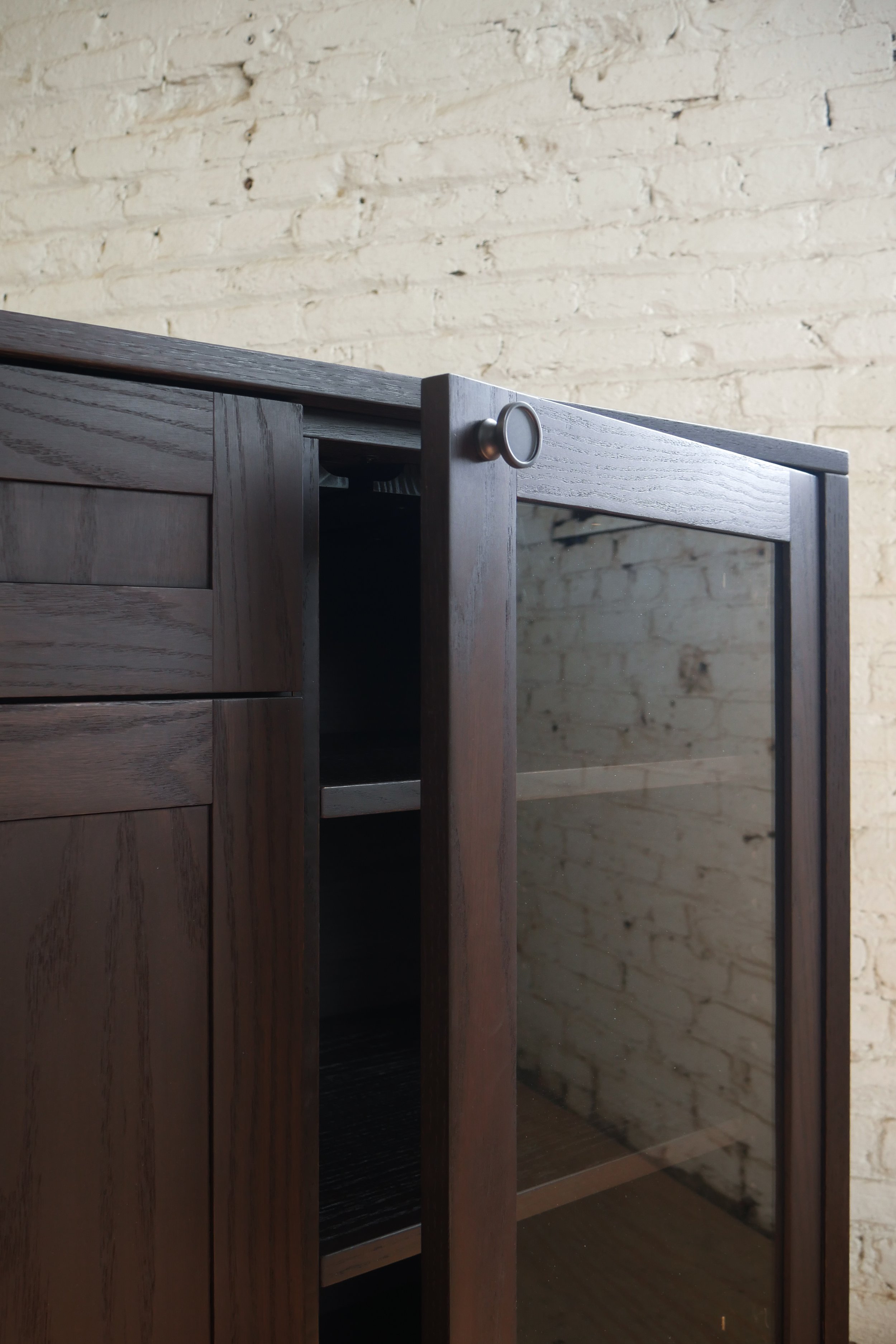 Close-up image of a wooden cabinet with a glass door and a round metal handle, set against a beige brick wall background.