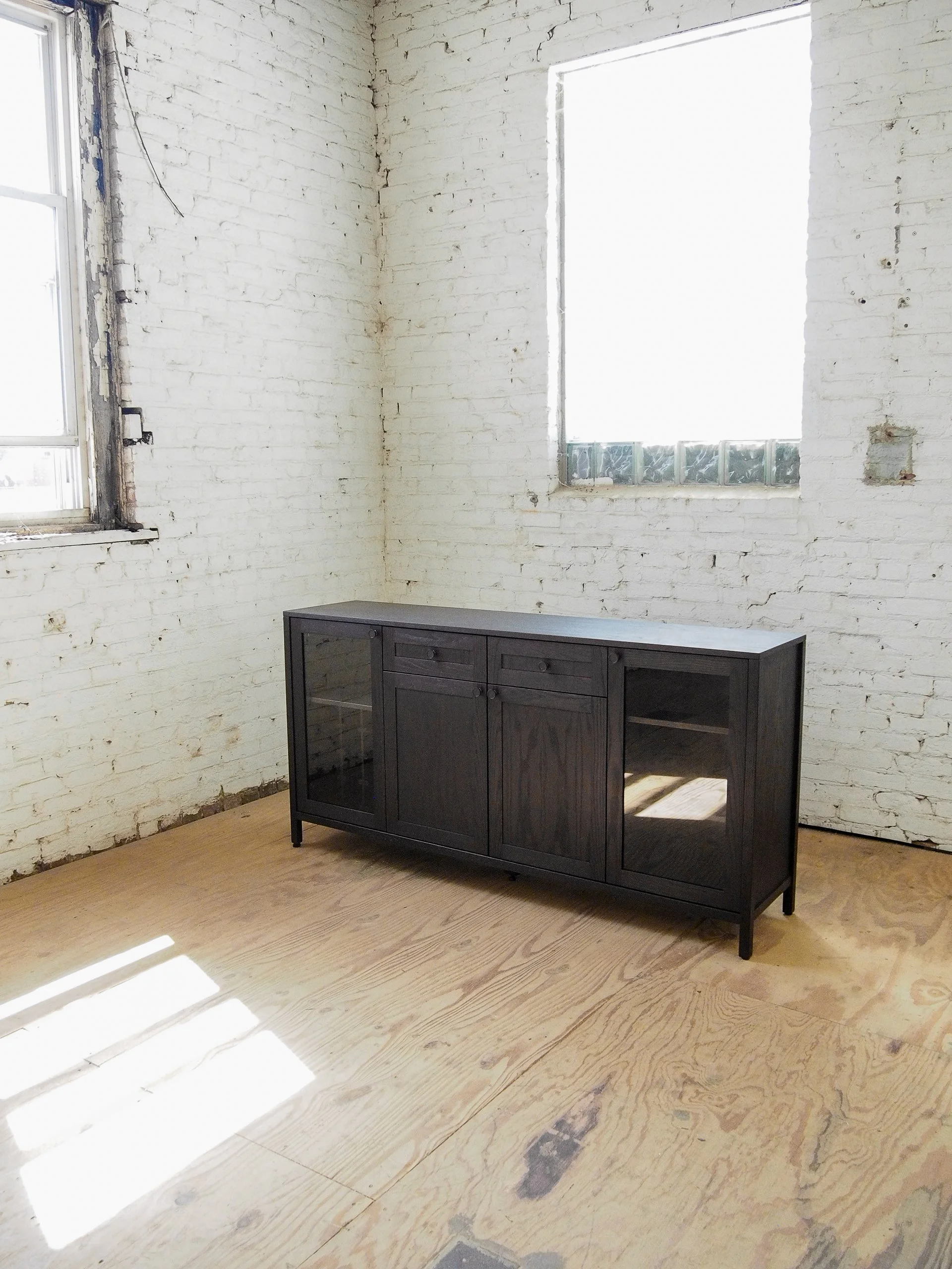 A room with white painted brick walls, two windows letting in bright sunlight, and a dark wooden cabinet on a light wooden floor.