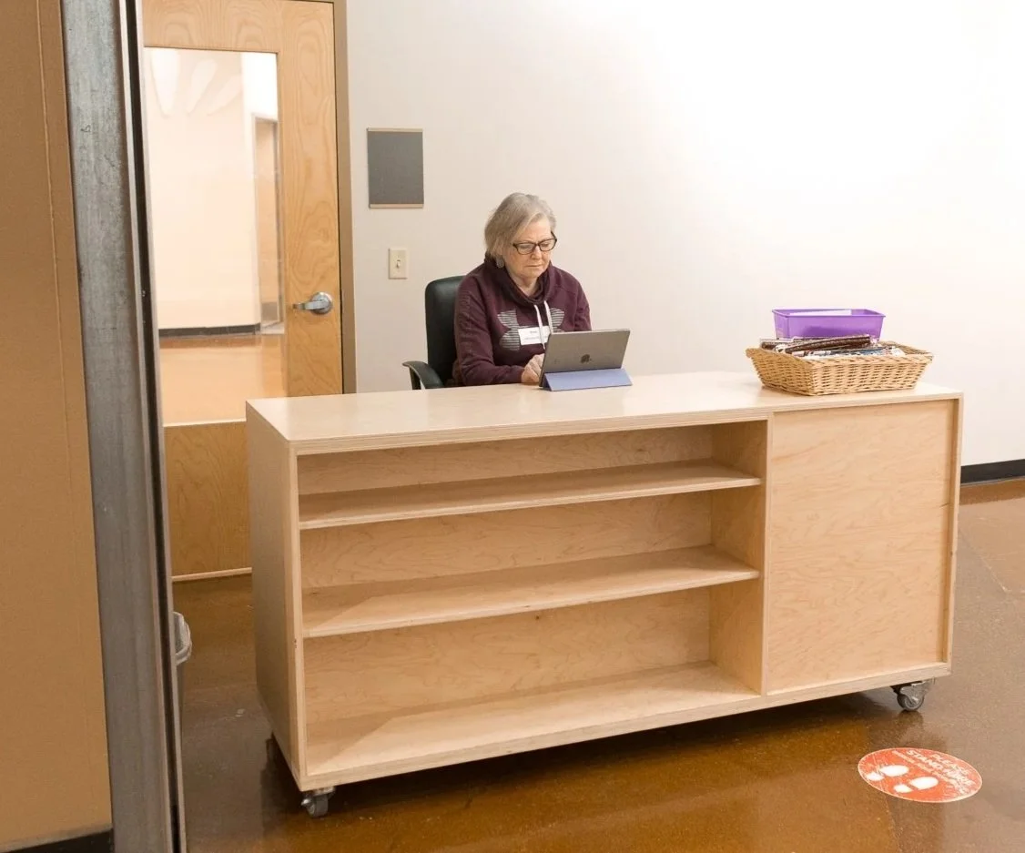 An elderly woman with glasses sitting at a custom wooden desk with a laptop in a room with plain white walls.