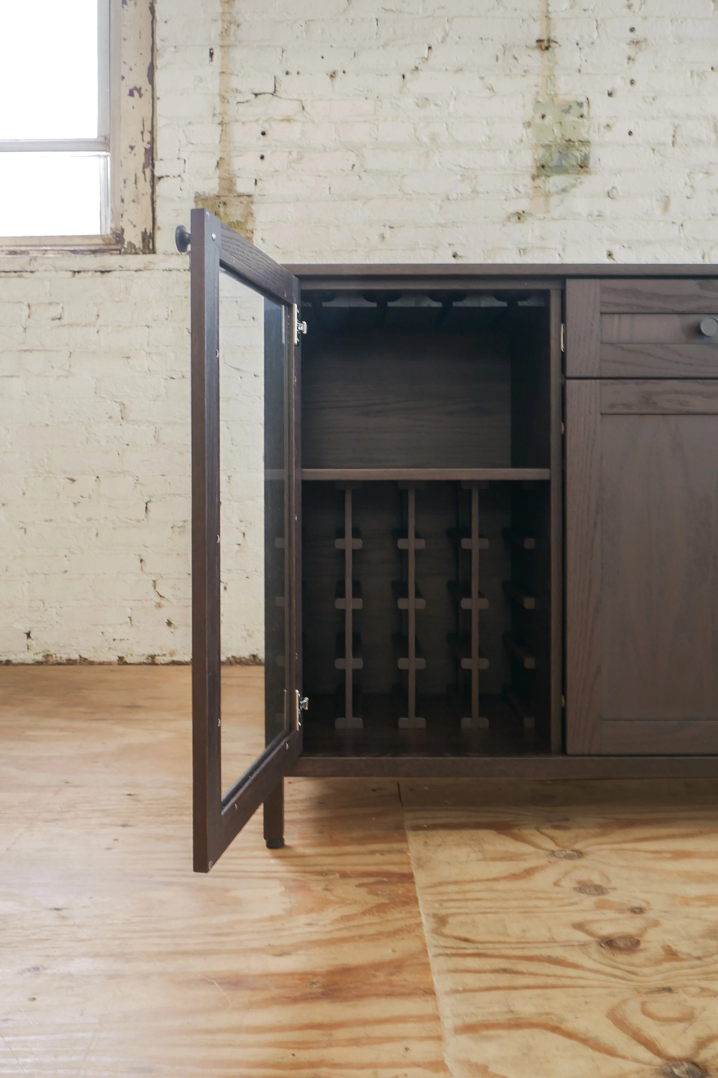 Open dark wooden cabinet with empty interior, set against a weathered brick wall with a small window in the upper left corner, and a wooden floor.