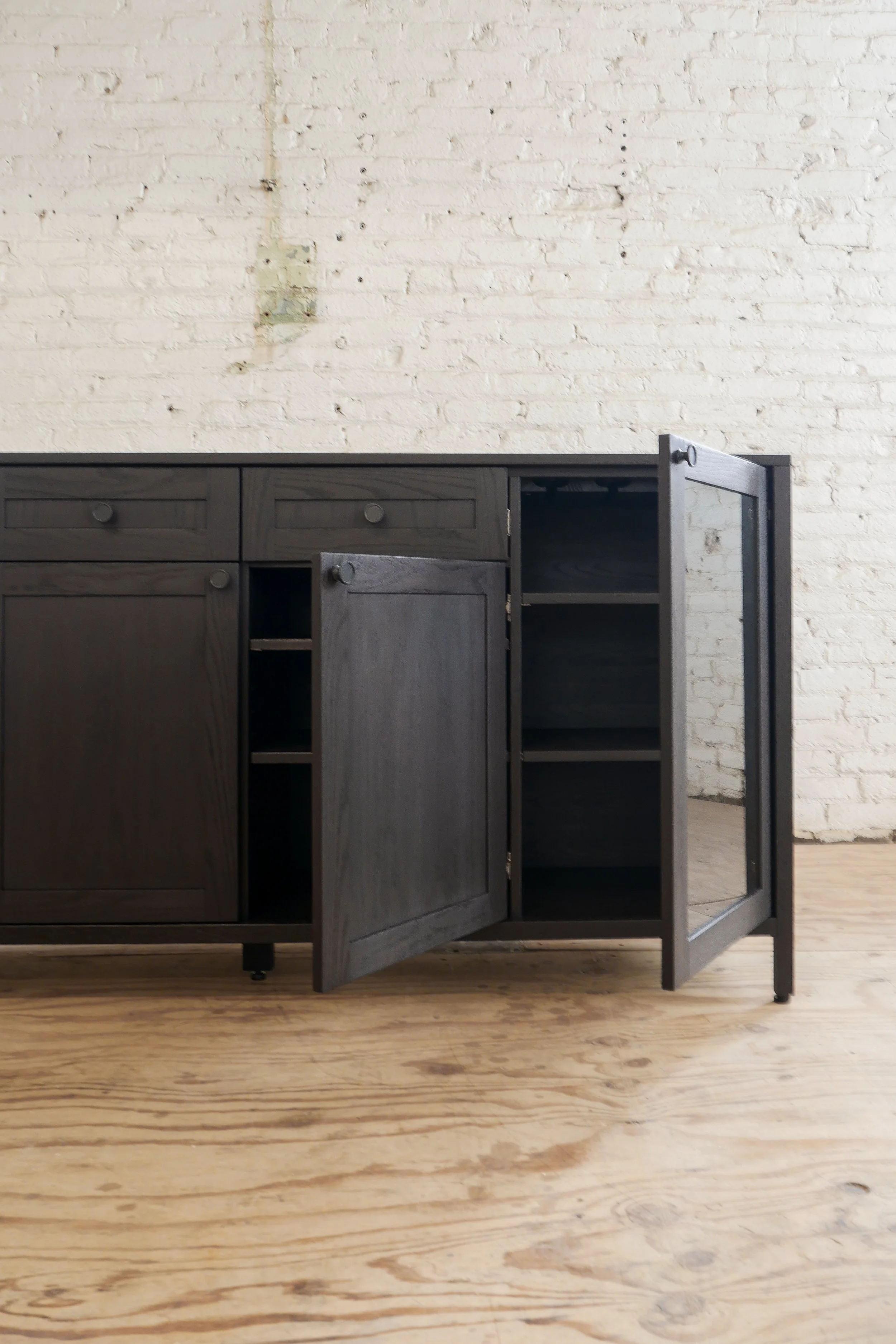 Empty black kitchen cabinet with open doors against a white brick wall on a wooden floor.